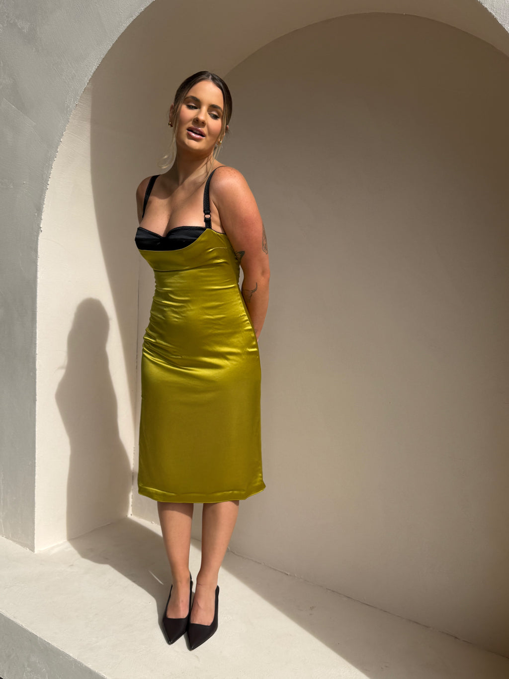 A woman modeling a shiny olive green midi dress with a black sweetheart neckline, standing in a minimalist white arched alcove.