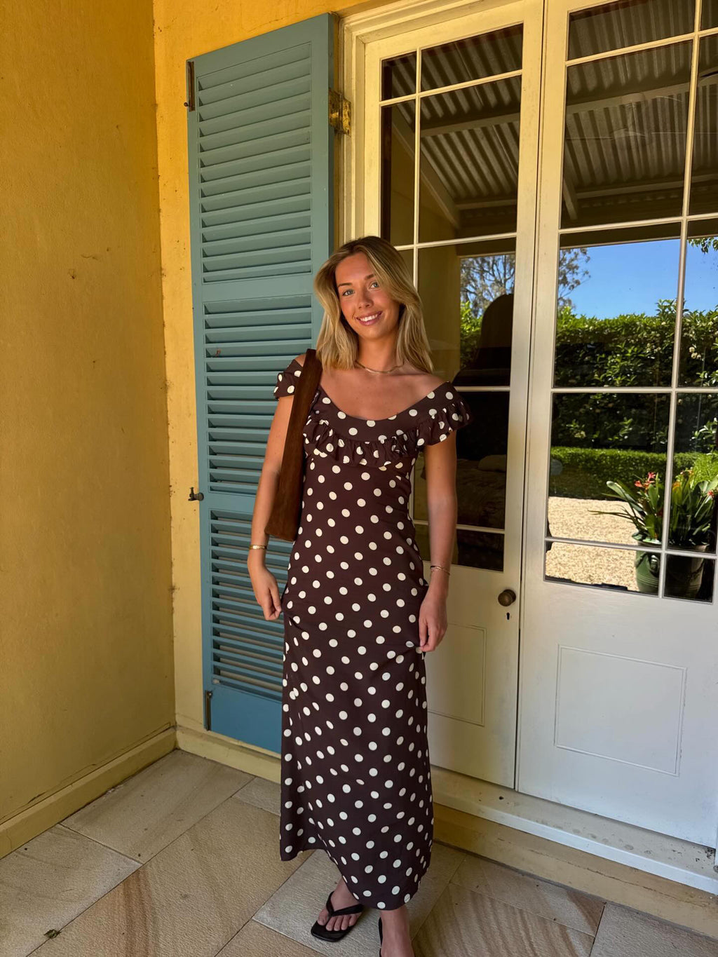A woman stands on a porch wearing a brown midi dress with white polka dots, a brown shoulder bag, and black sandals, posing in front of a yellow wall, blue shutters, and white glass doors. - @emmakelaher