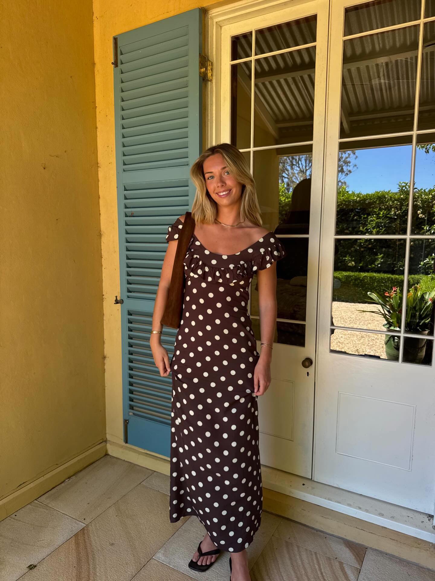 A woman stands on a porch wearing a brown midi dress with white polka dots, a brown shoulder bag, and black sandals, posing in front of a yellow wall, blue shutters, and white glass doors. - @emmakelaher