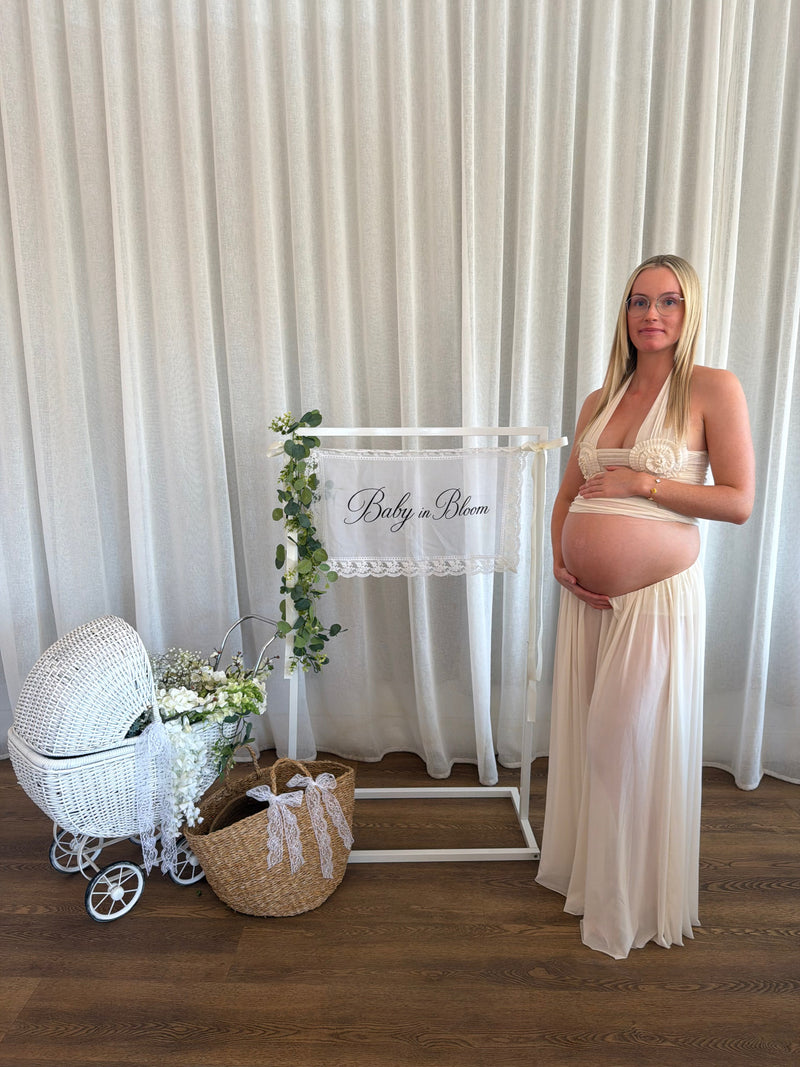 A pregnant woman stands in a studio wearing a cream halter-neck maternity gown with a matching bodice and flowing skirt, holding her baby bump next to a white sign that reads 'Baby in Bloom'. A white wicker bassinet, a woven basket with white ribbons, and greenery are arranged on a wooden floor against a white curtain backdrop. - @ashley_mariee