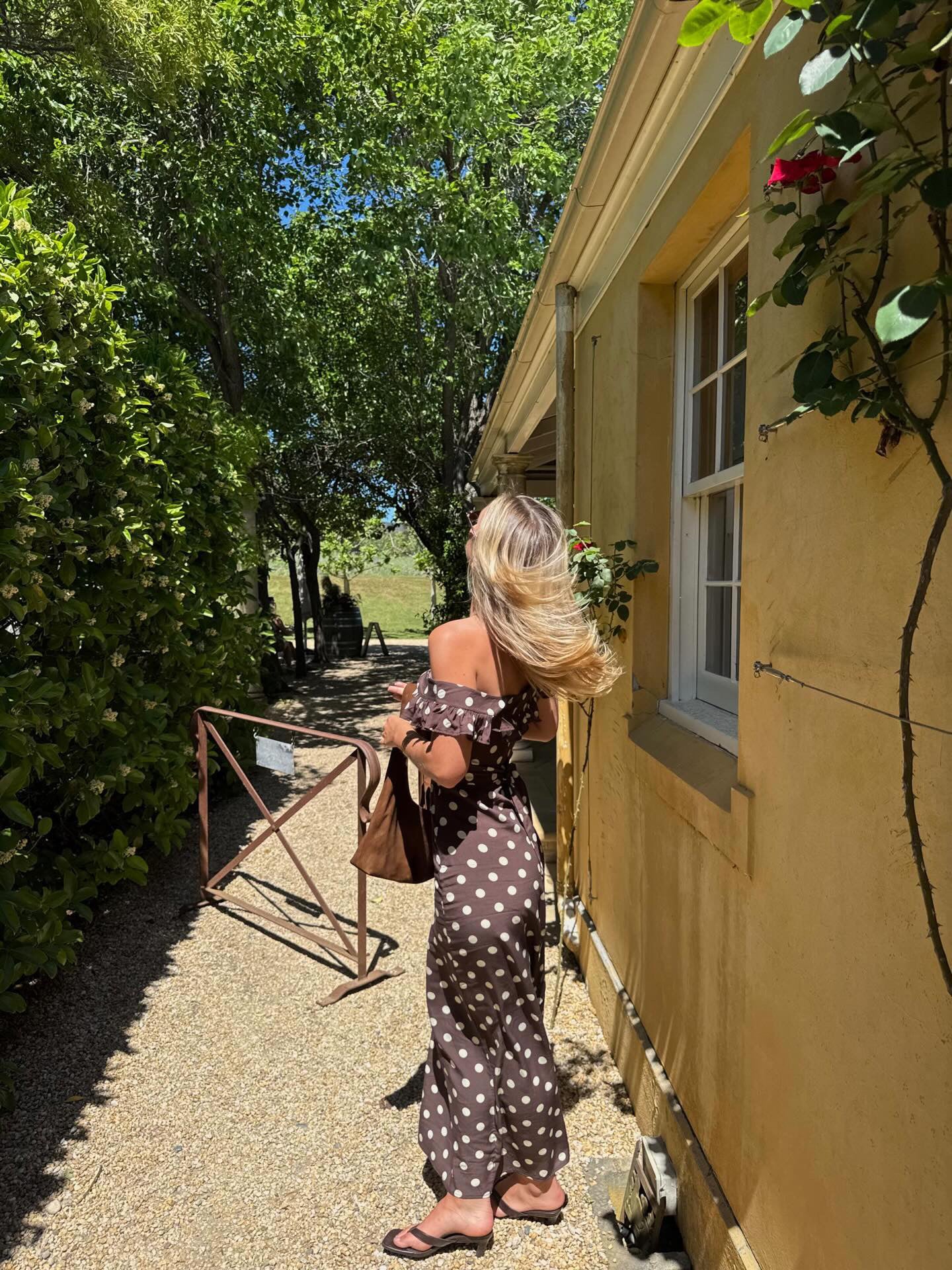 A woman stands on a gravel path beside a yellow stucco building, wearing an off-the-shoulder brown maxi dress with white polka dots and carrying a brown tote bag.