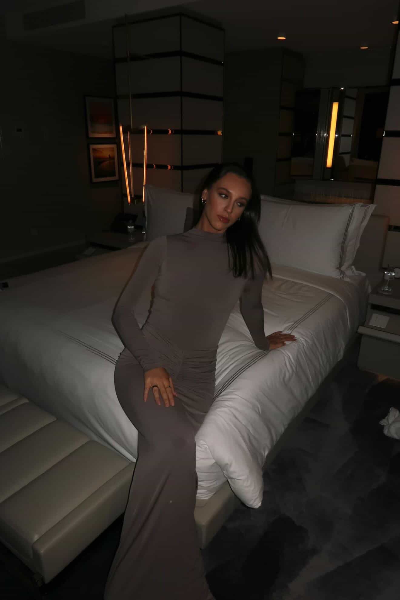 Woman in a gray dress sitting on a bed in a dimly lit room.