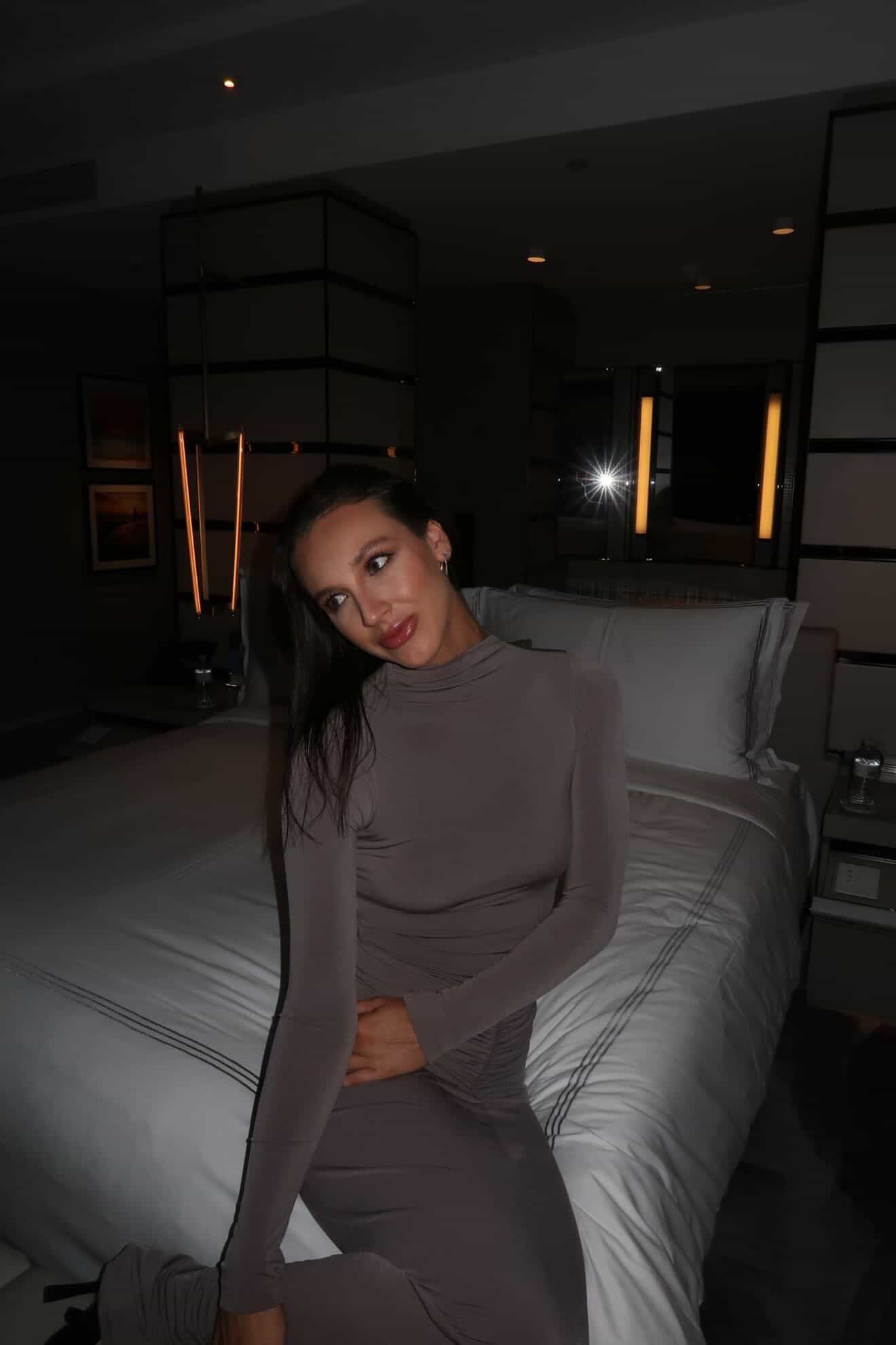 Woman in a gray outfit sitting on a bed in a dimly lit room.