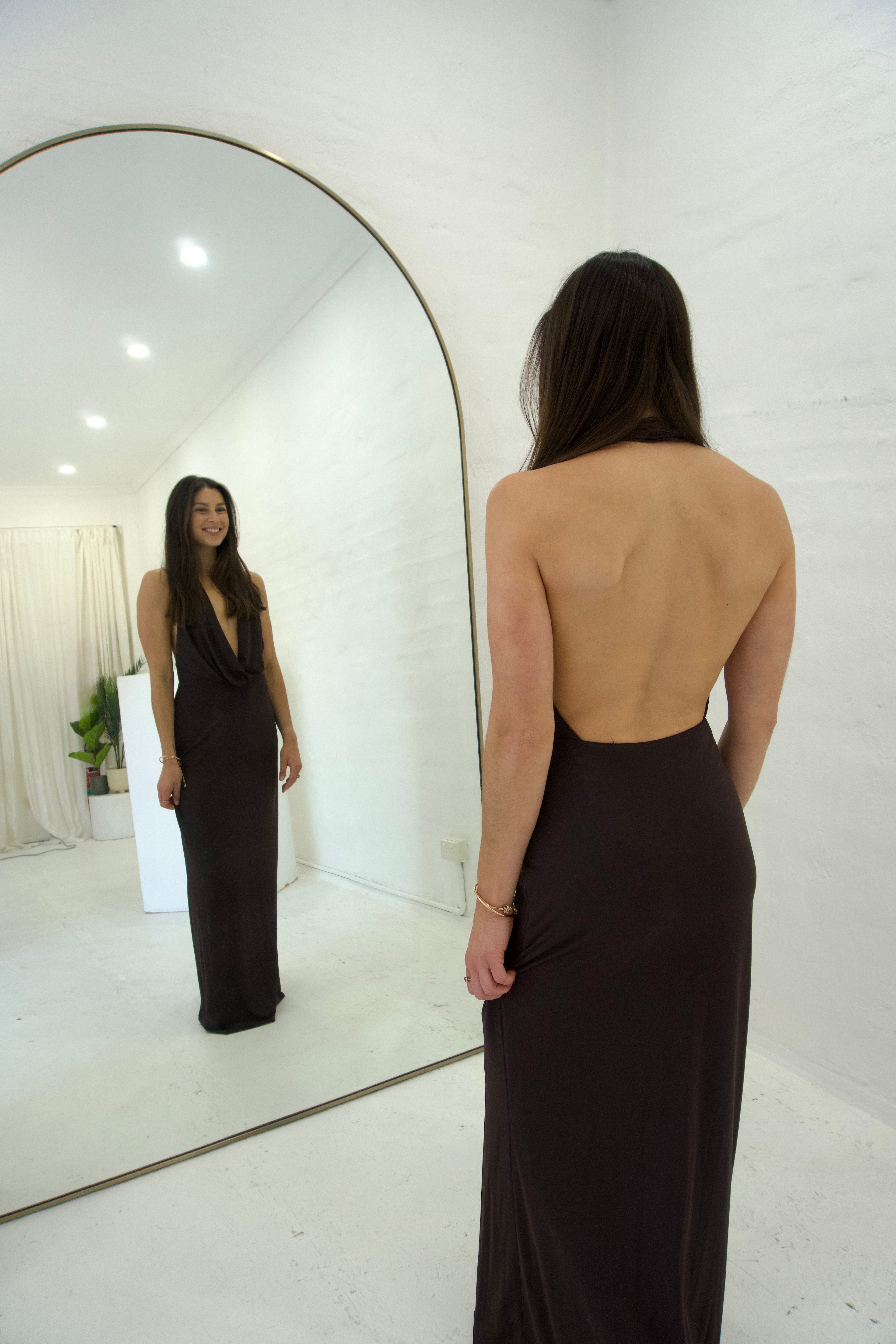 Woman in a black evening gown standing in front of a mirror in a minimalistic room.