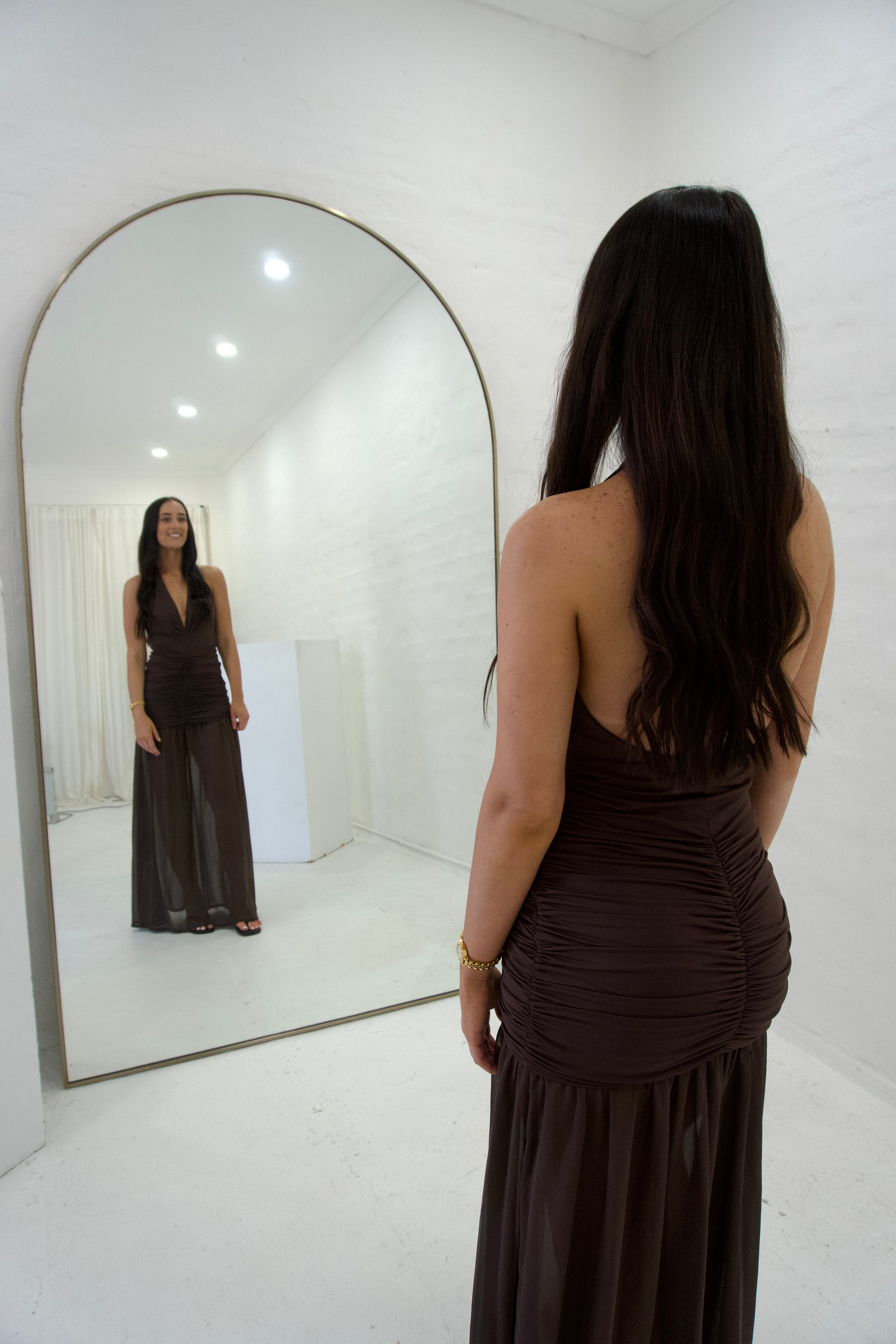 Woman in a long dark dress standing in front of a large mirror in a minimalistic room.