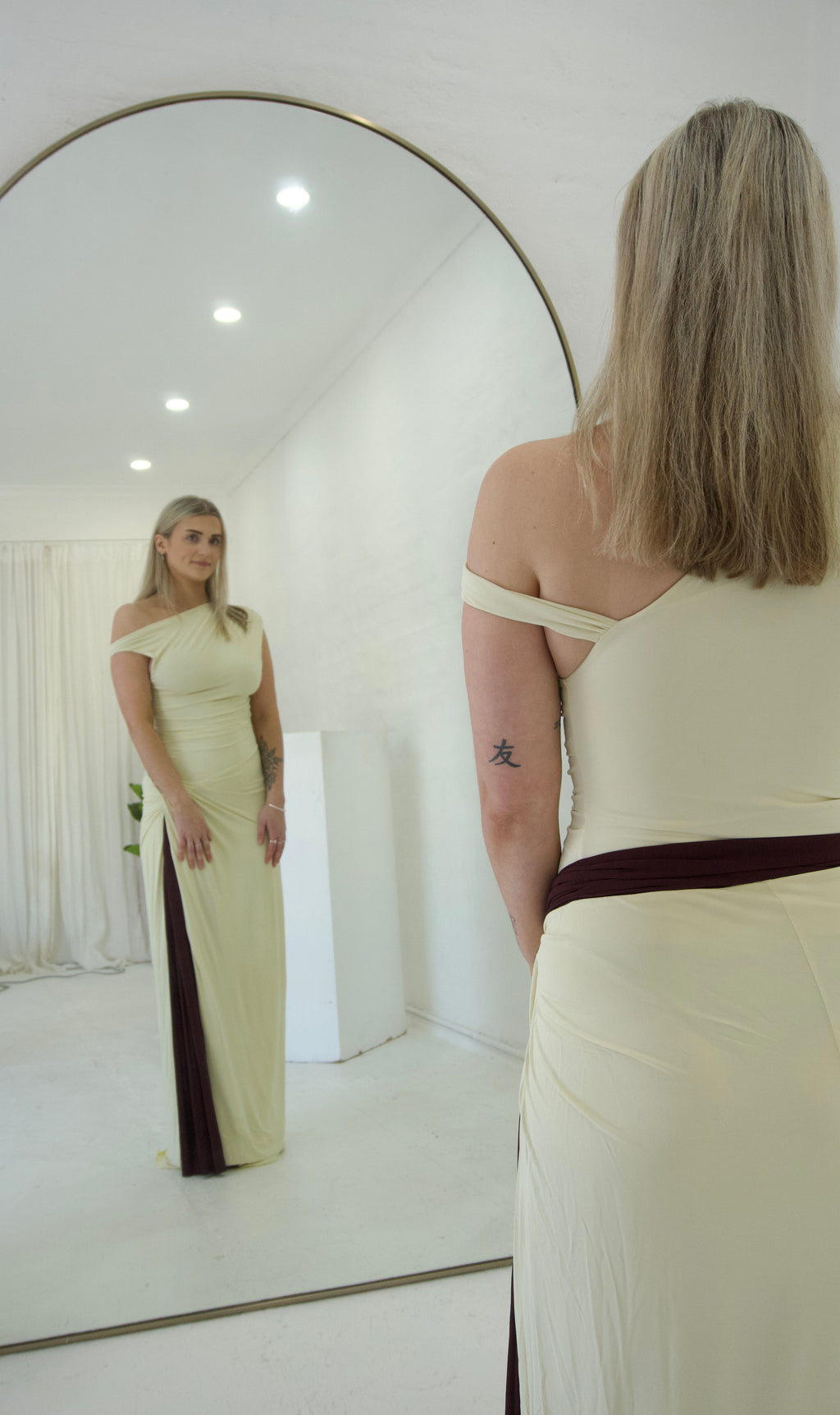 Woman in a long off-shoulder dress standing in front of a large round mirror.
