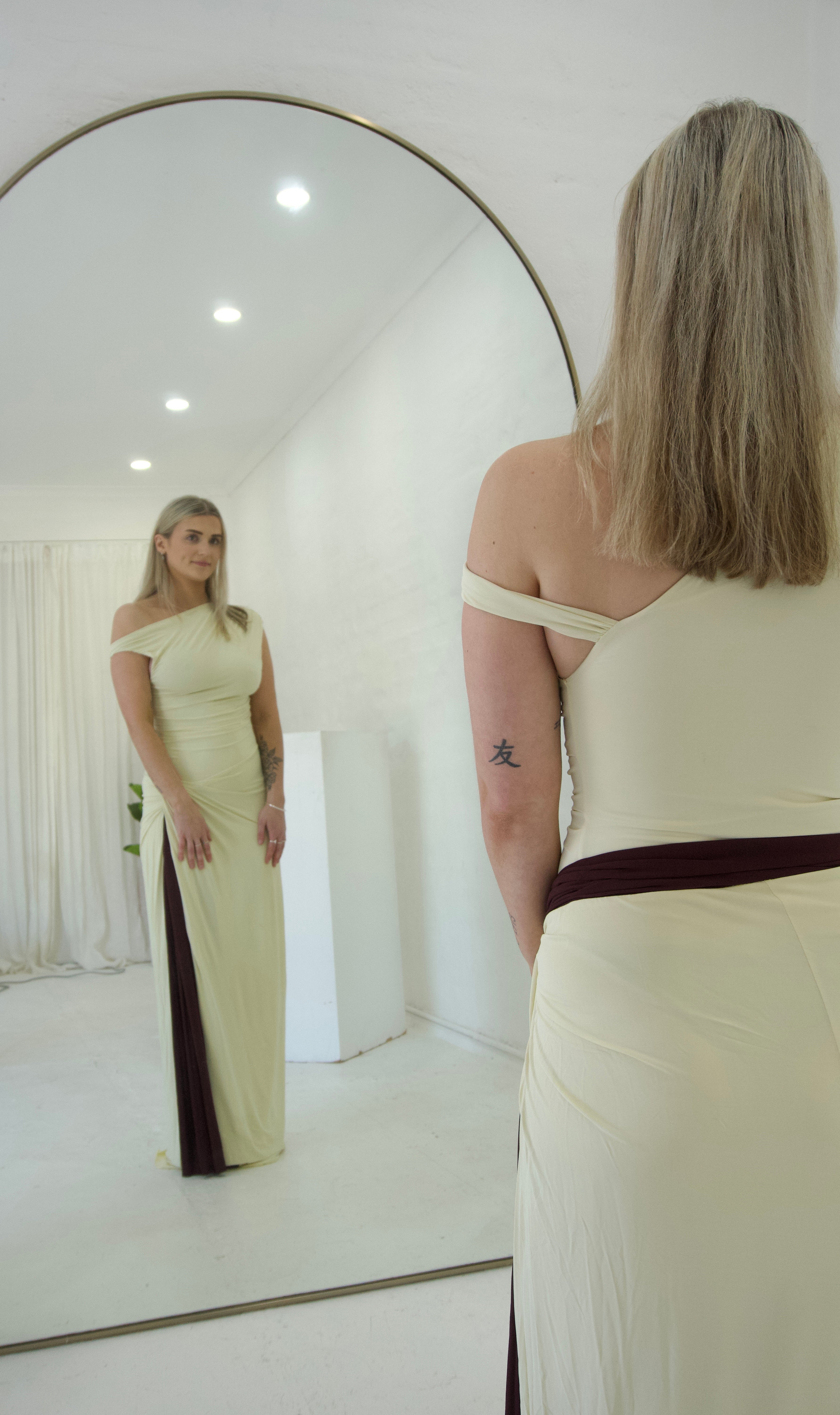 Woman in a long off-shoulder dress standing in front of a large round mirror.