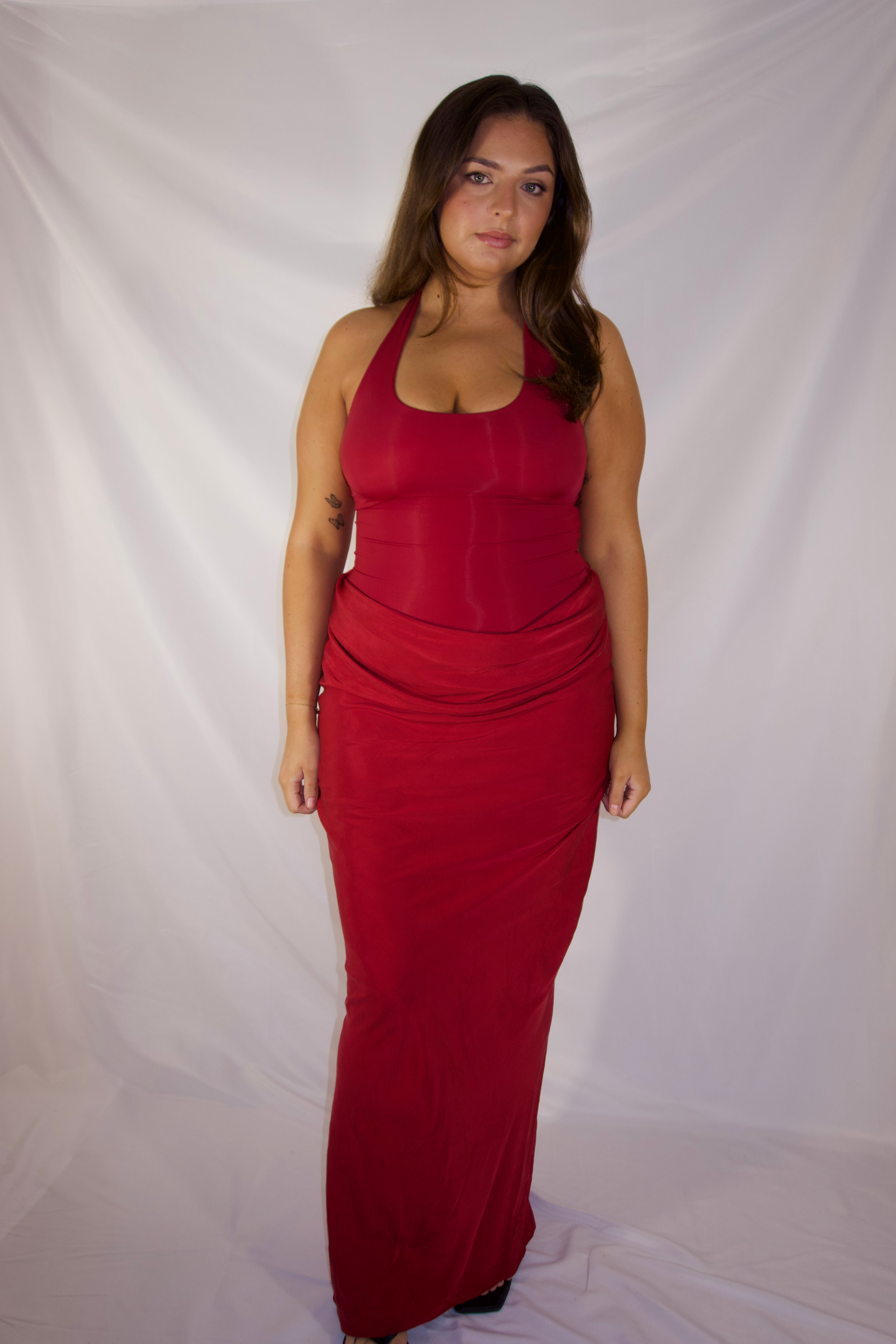 A woman modeling a sleeveless, floor-length red dress against a white fabric backdrop.