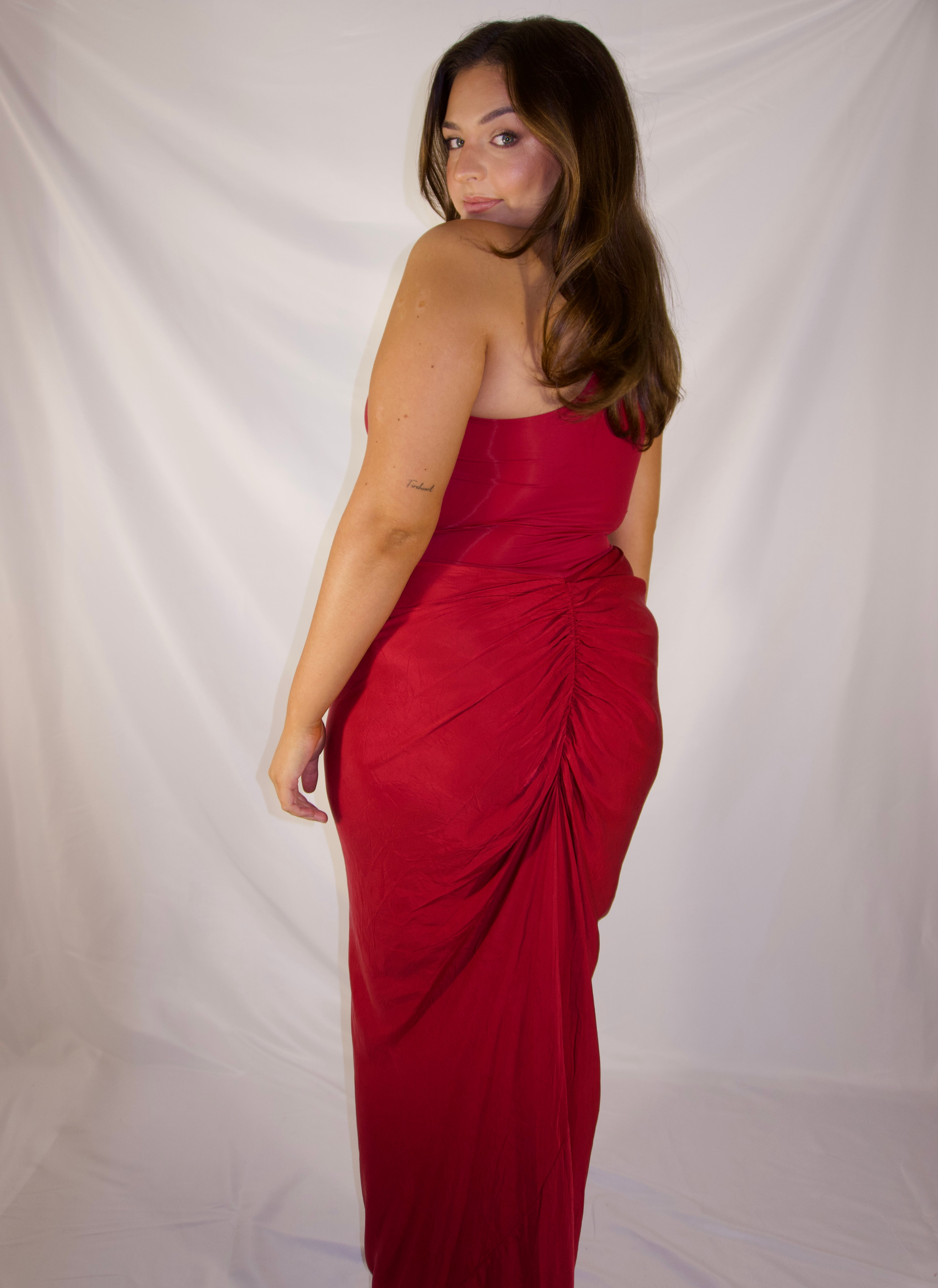 A woman modeling a strapless red maxi dress with a gathered ruched skirt, photographed against a white fabric backdrop.