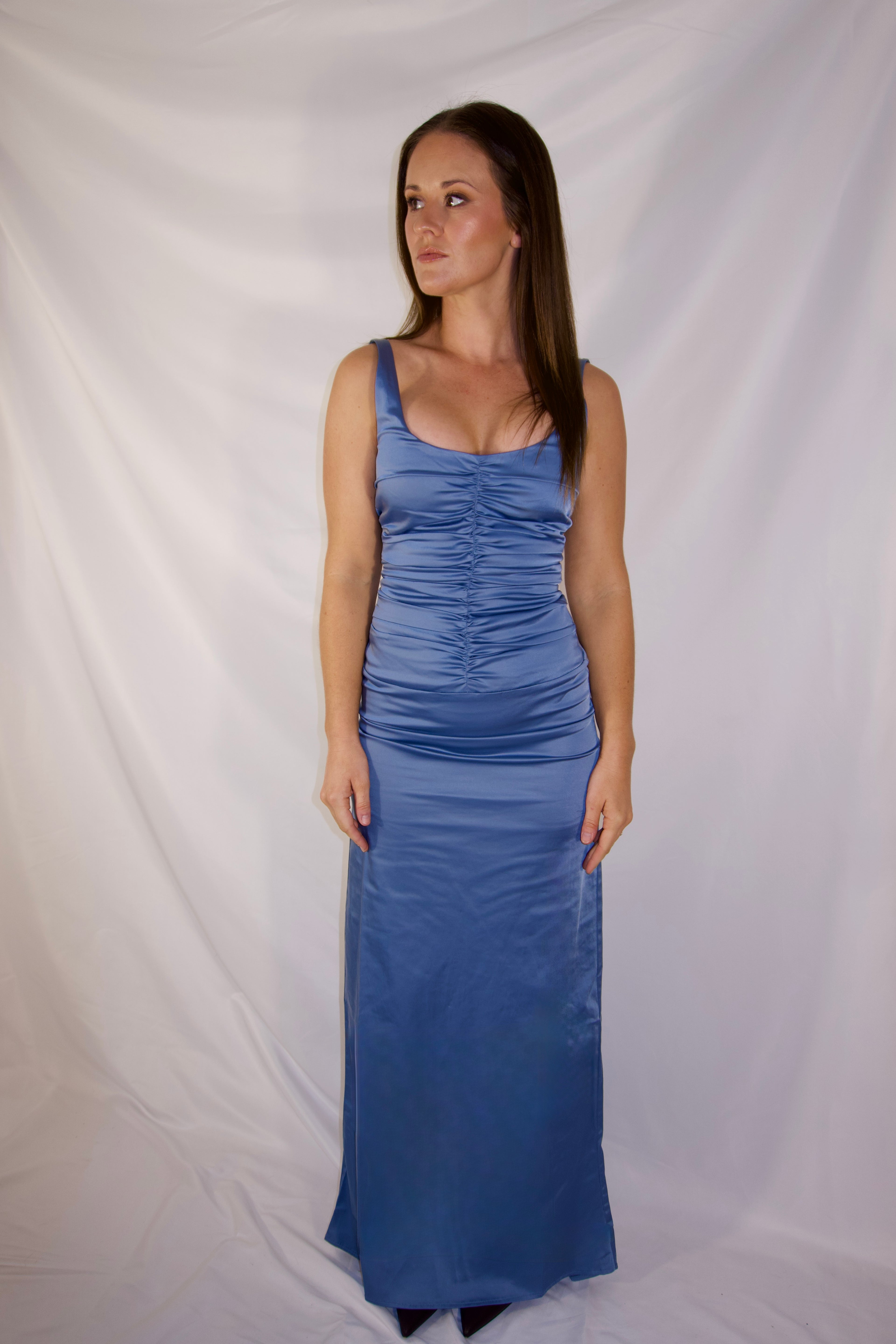 A woman modeling a sleeveless, floor-length blue satin dress with a ruched bodice, standing against a draped white fabric backdrop.