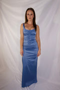 A woman modeling a sleeveless, floor-length blue satin gown with ruched detailing, standing against a draped white fabric backdrop.