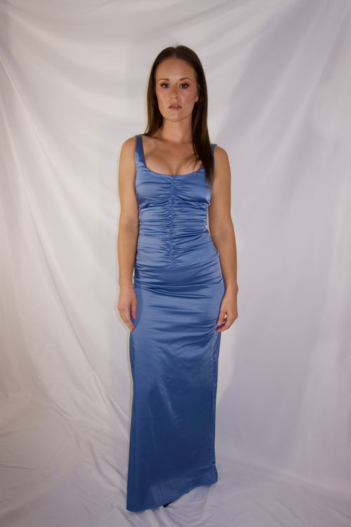 A woman modeling a sleeveless, floor-length blue satin gown with ruched detailing, standing against a draped white fabric backdrop.