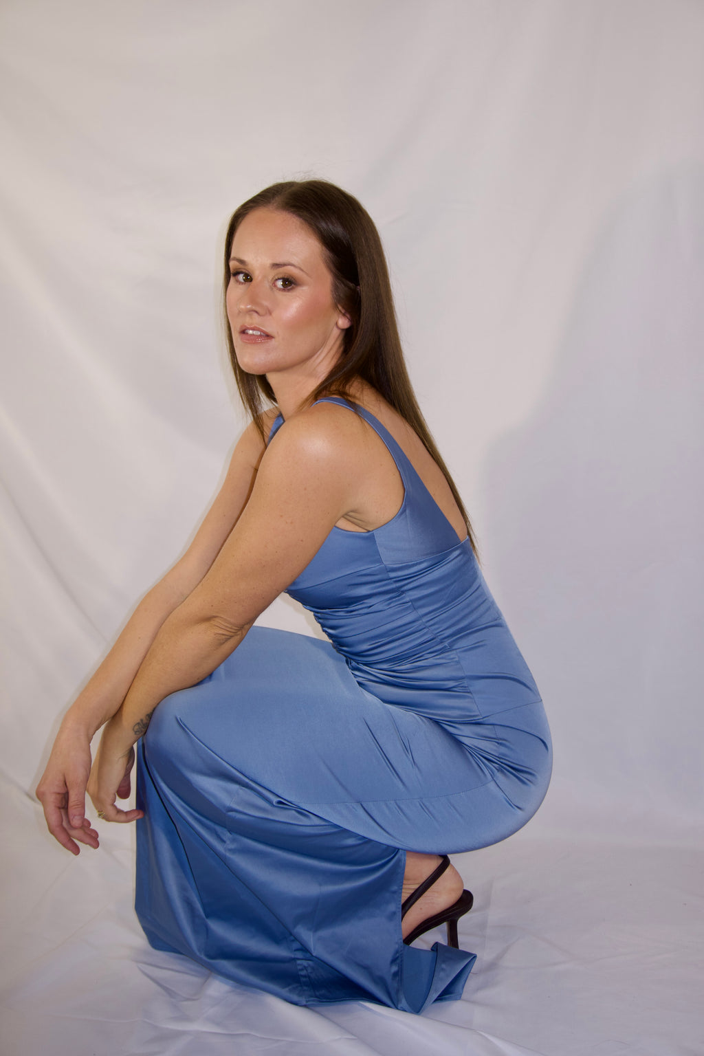 A woman wearing a blue satin maxi dress and black strappy heels, crouching in a studio setting against a white fabric backdrop.