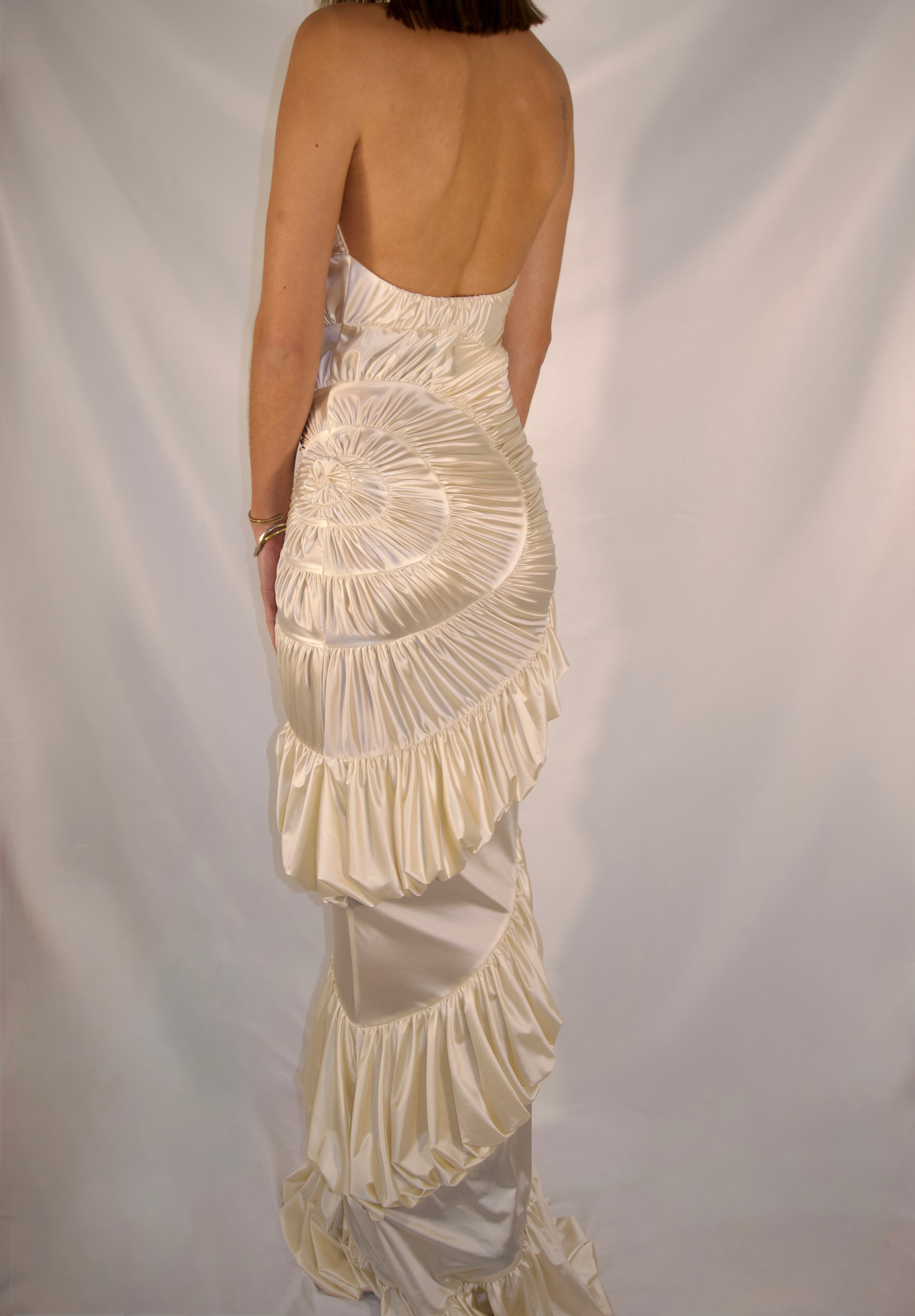 A woman shown from the back wearing a strapless, open-back dress with dramatic gathered ruffles and layered tiers in a warm ivory or cream tone, photographed against a white fabric backdrop.