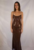 A woman modeling a floor-length, spaghetti-strap brown sequined dress against a white fabric backdrop. - @_ellahabates
