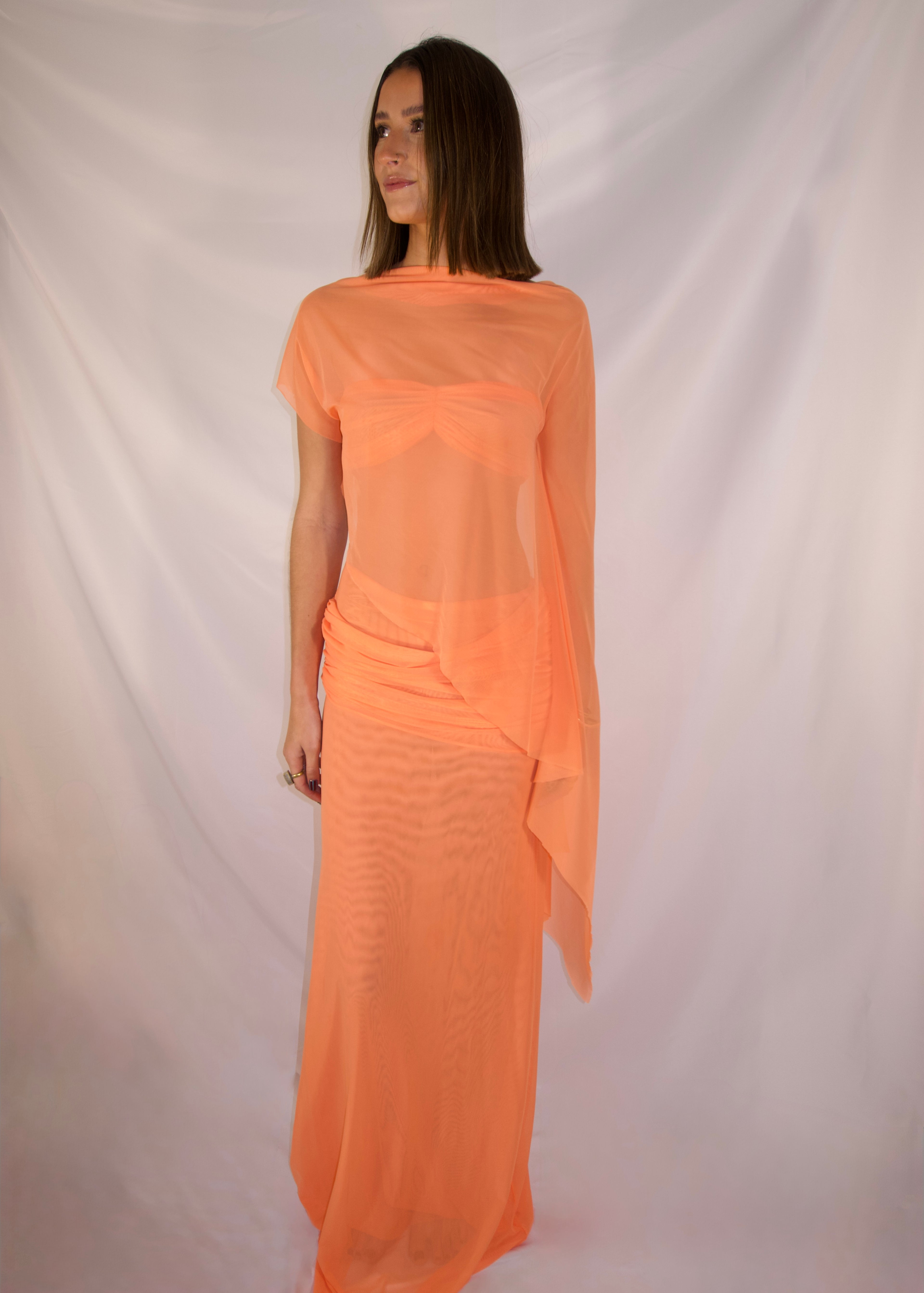 A woman modeling a sheer peach-orange maxi dress with short sleeves and a draped, flowing overlay, photographed against a white fabric backdrop. - @_ellahbates