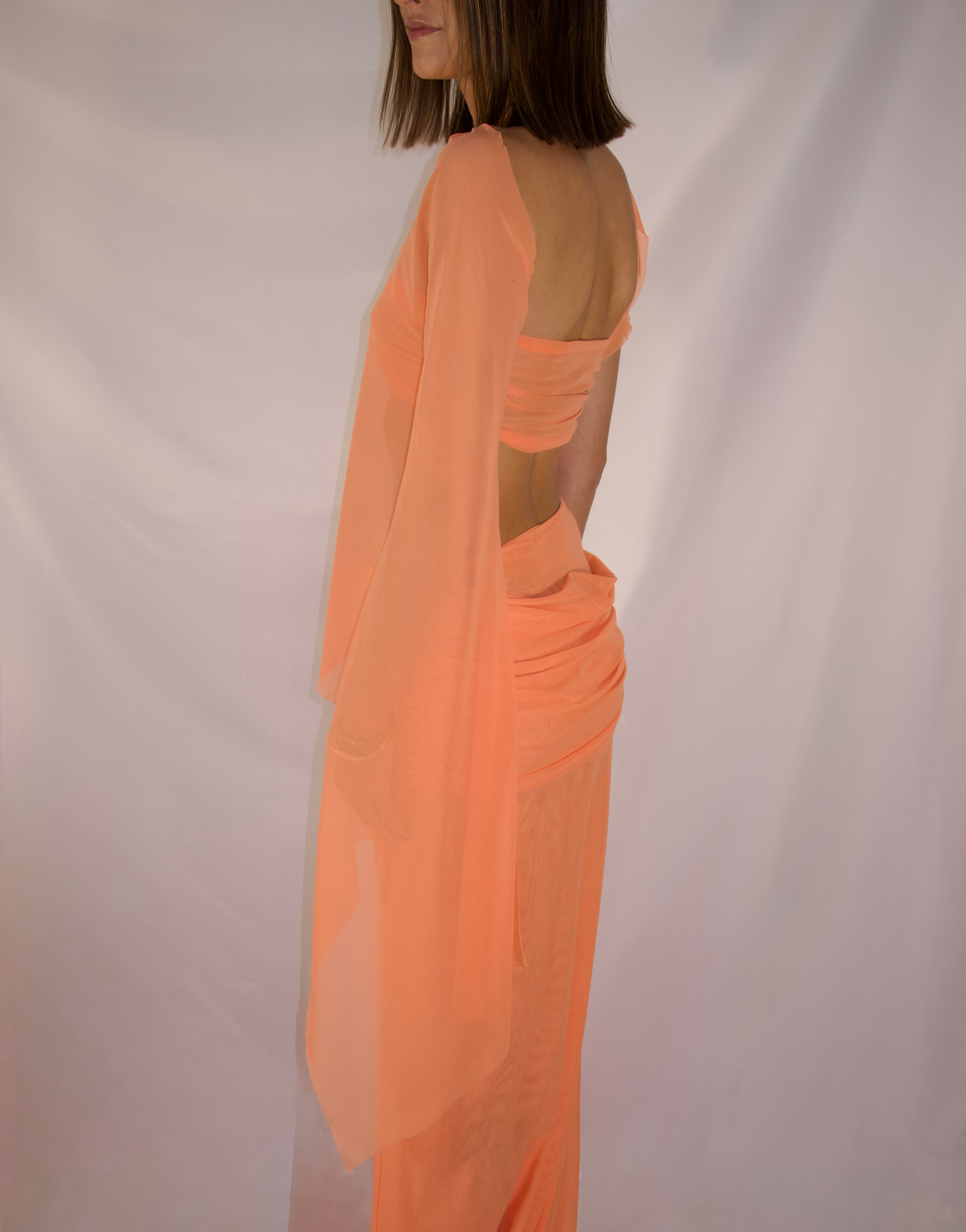 A woman shown from the back wearing a sheer peach-orange draped dress or sarong with a matching draped wrap, photographed against a plain light backdrop.