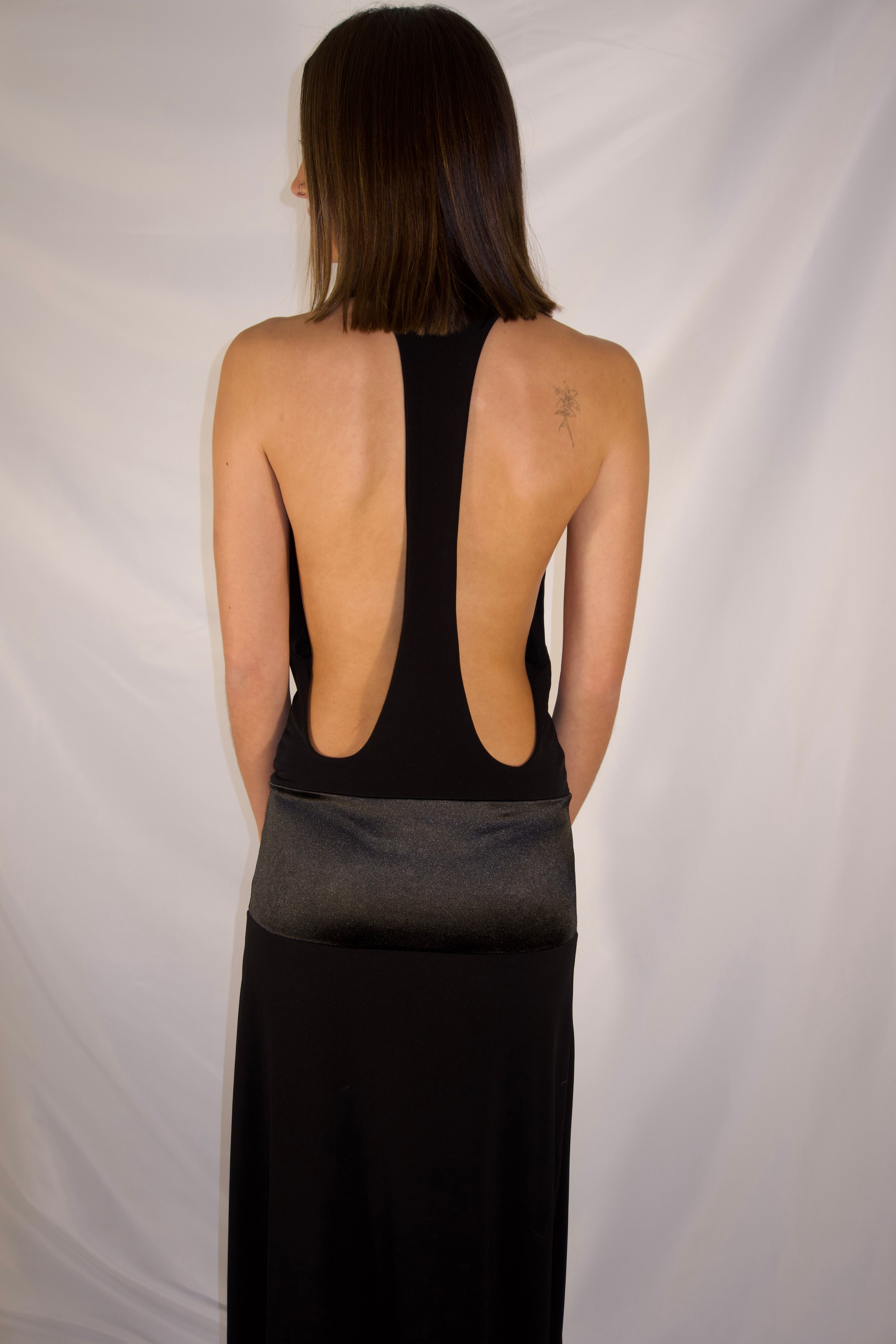 A woman shown from the back wearing a black open-back dress with a sheer smoky gray waistband, photographed against a white fabric backdrop.