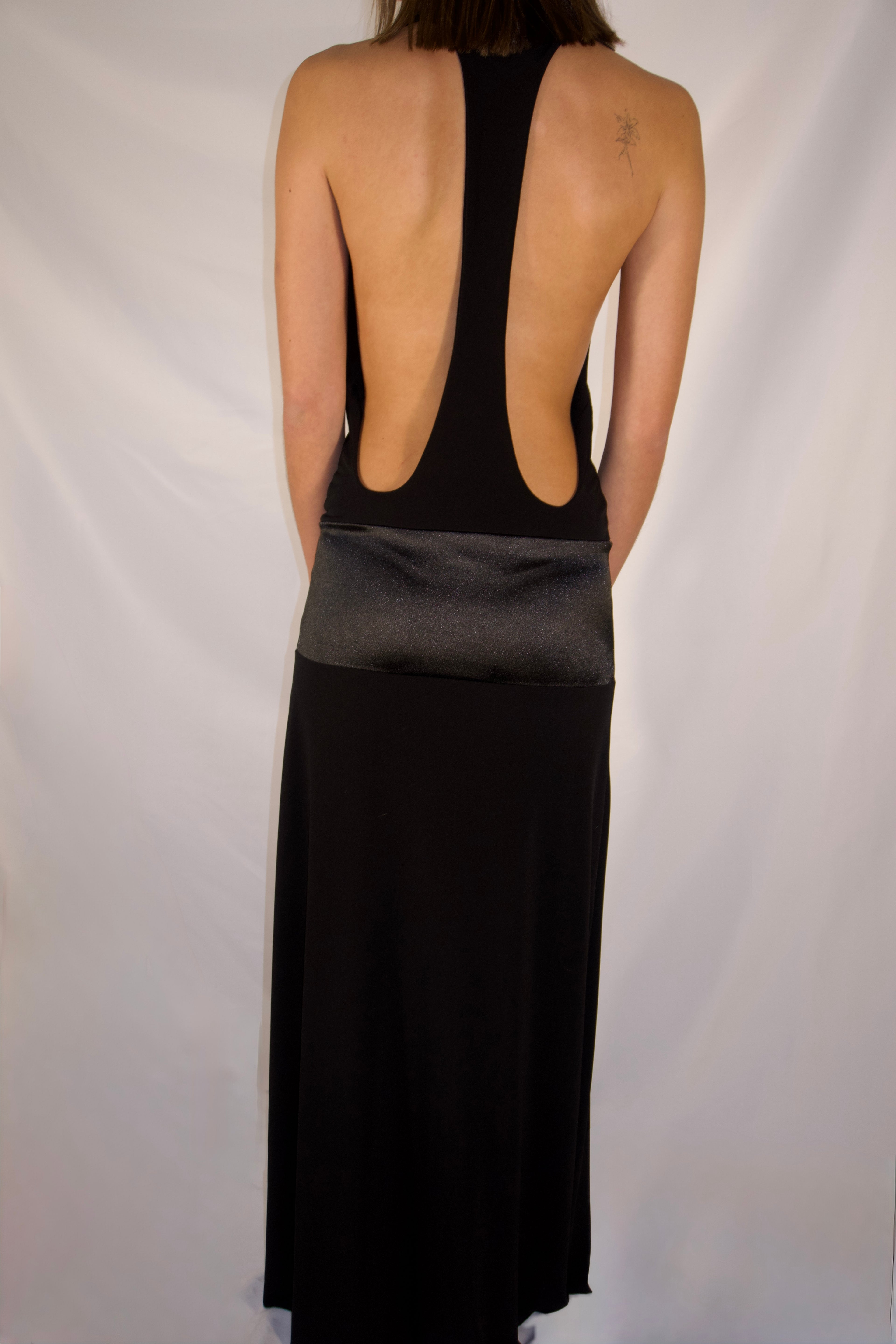A woman shown from the back wearing a black maxi dress with a wide open back and a satin-like waistband, photographed against a white studio backdrop.