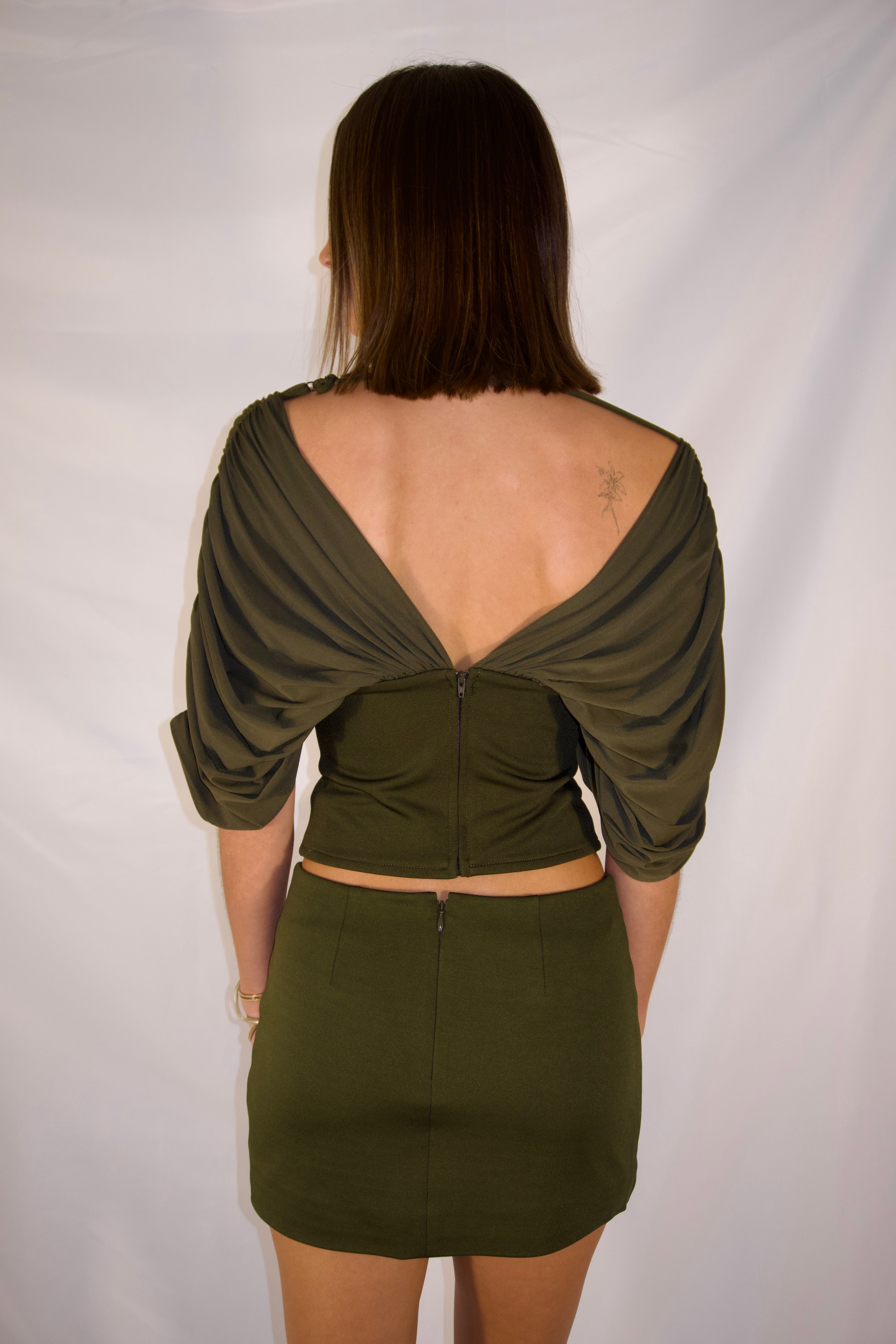 A woman shown from the back wearing an olive green two-piece outfit consisting of a ruched crop top with draped sleeves and a matching mini skirt, set against a white studio backdrop.