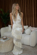 A woman models a sleeveless, pleated cream-colored gown with a halter neckline and dramatic cascading ruffles, standing in a modern living room setting with a white sofa and brown curtains.