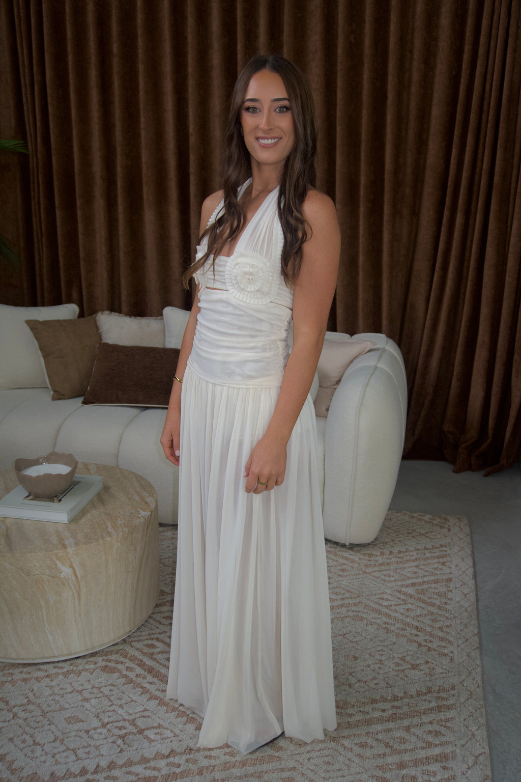 A woman wearing a sleeveless white halter-neck maxi dress with a textured bodice and flowing skirt, standing in a living room with brown curtains, a cream sofa, and a patterned rug.