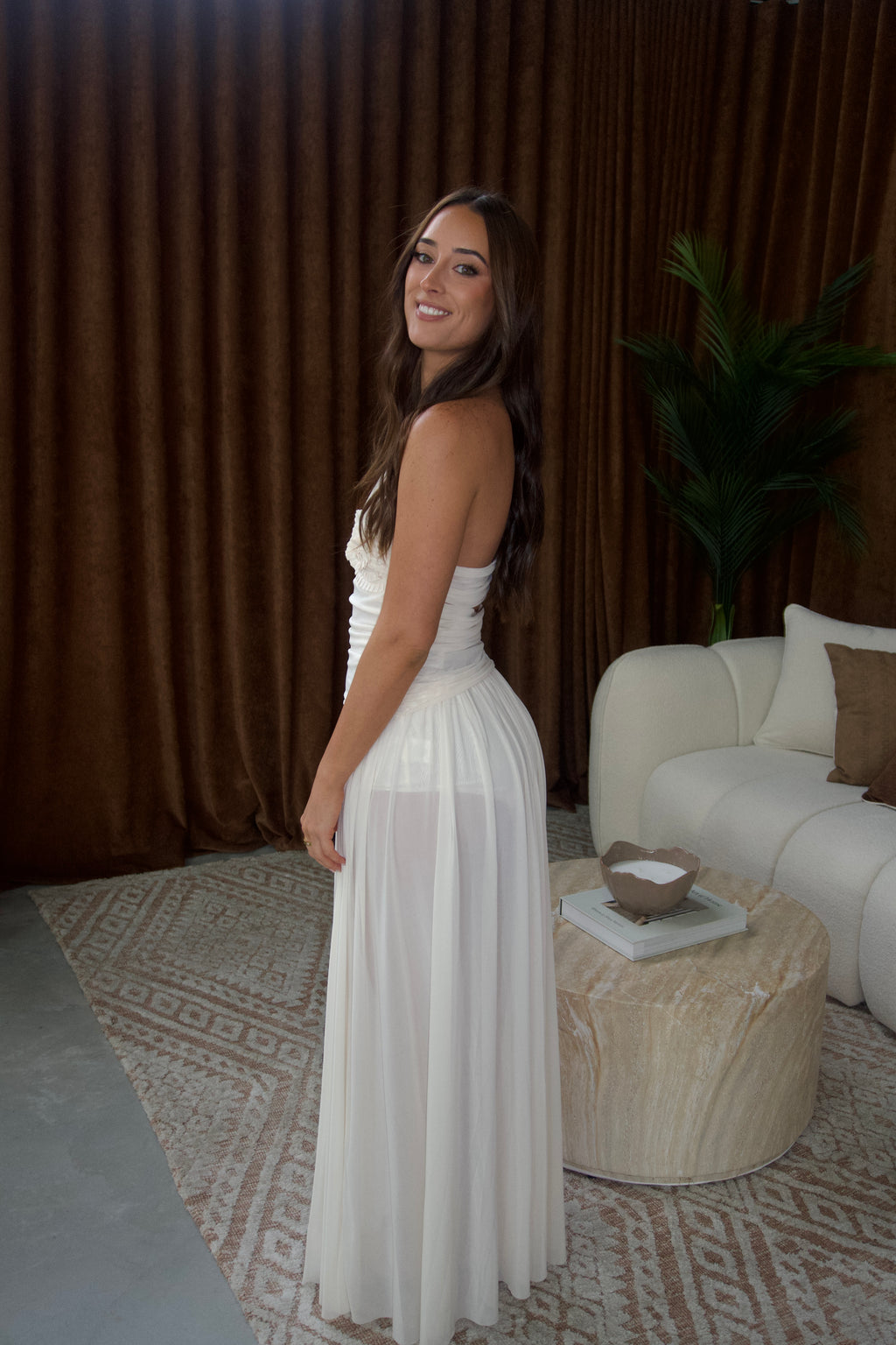 A woman wearing a strapless white maxi dress stands in a styled room with brown curtains, a white sofa, and a patterned rug.