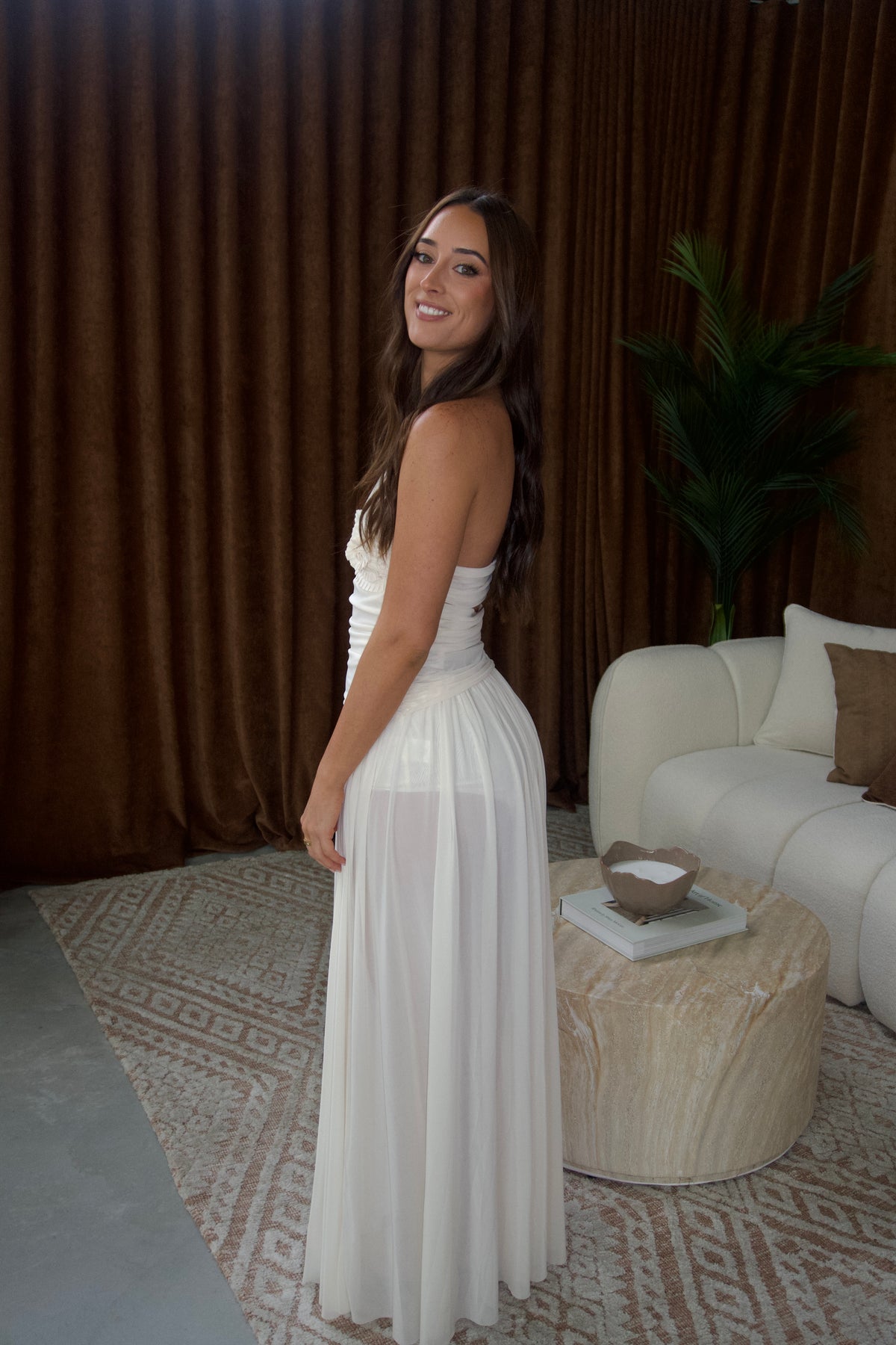A woman wearing a strapless white maxi dress stands in a styled room with brown curtains, a white sofa, and a patterned rug.