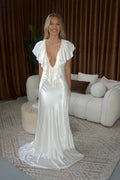 A woman wearing a floor-length white satin gown with ruffled short sleeves and a deep plunging neckline, standing in a living room with brown curtains, a cream sofa, and a patterned rug. - @indiabluegranger