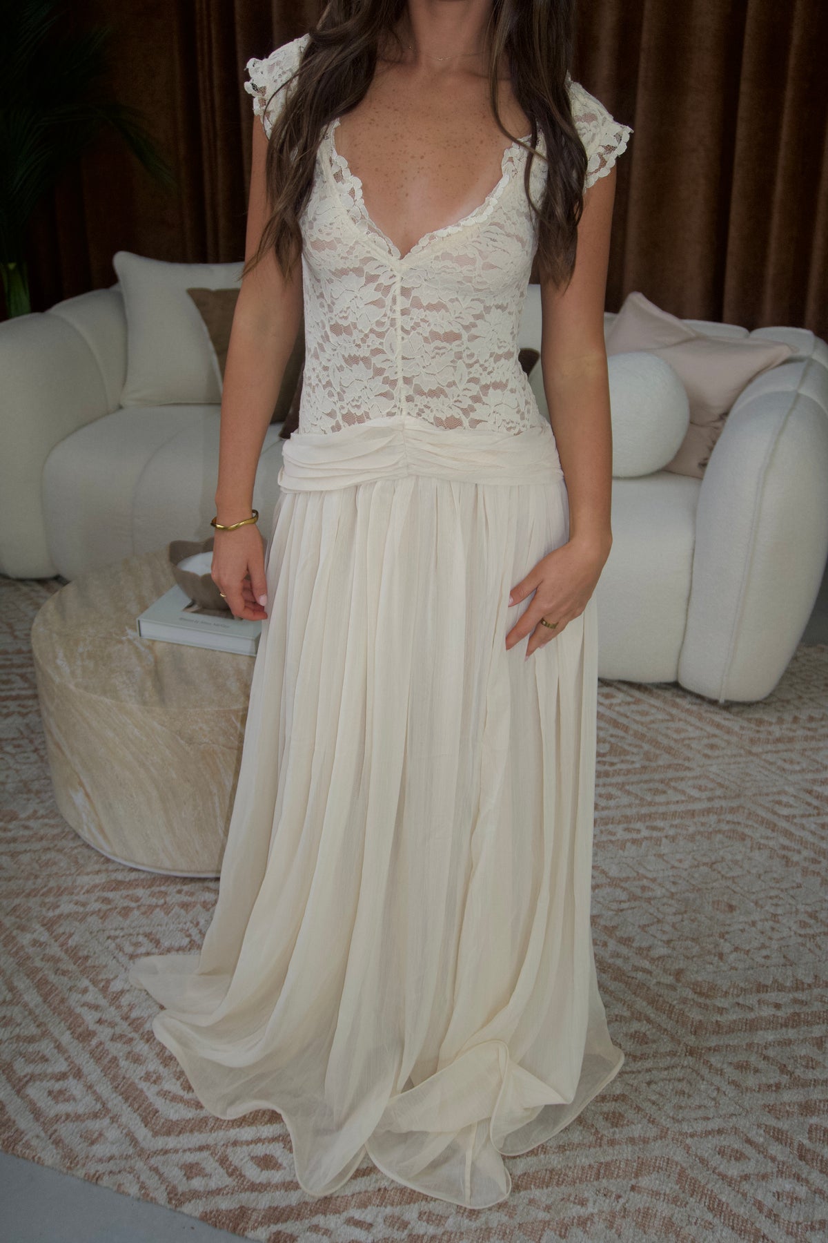 Woman wearing an ivory floor-length gown featuring a fitted floral lace bodice with cap sleeves, plunging neckline and a flowing pleated skirt with ruched waist detailing, photographed in a neutral studio setting.