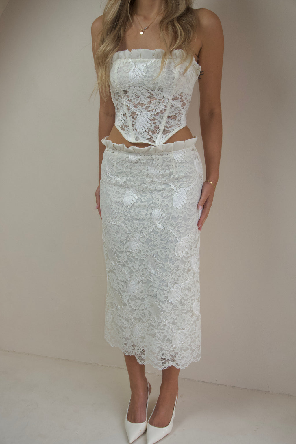  Woman wearing an ivory lace two-piece set featuring a strapless corset-style top with ruffle detailing and a matching lace midi skirt, styled with white pointed heels against a neutral studio background.
