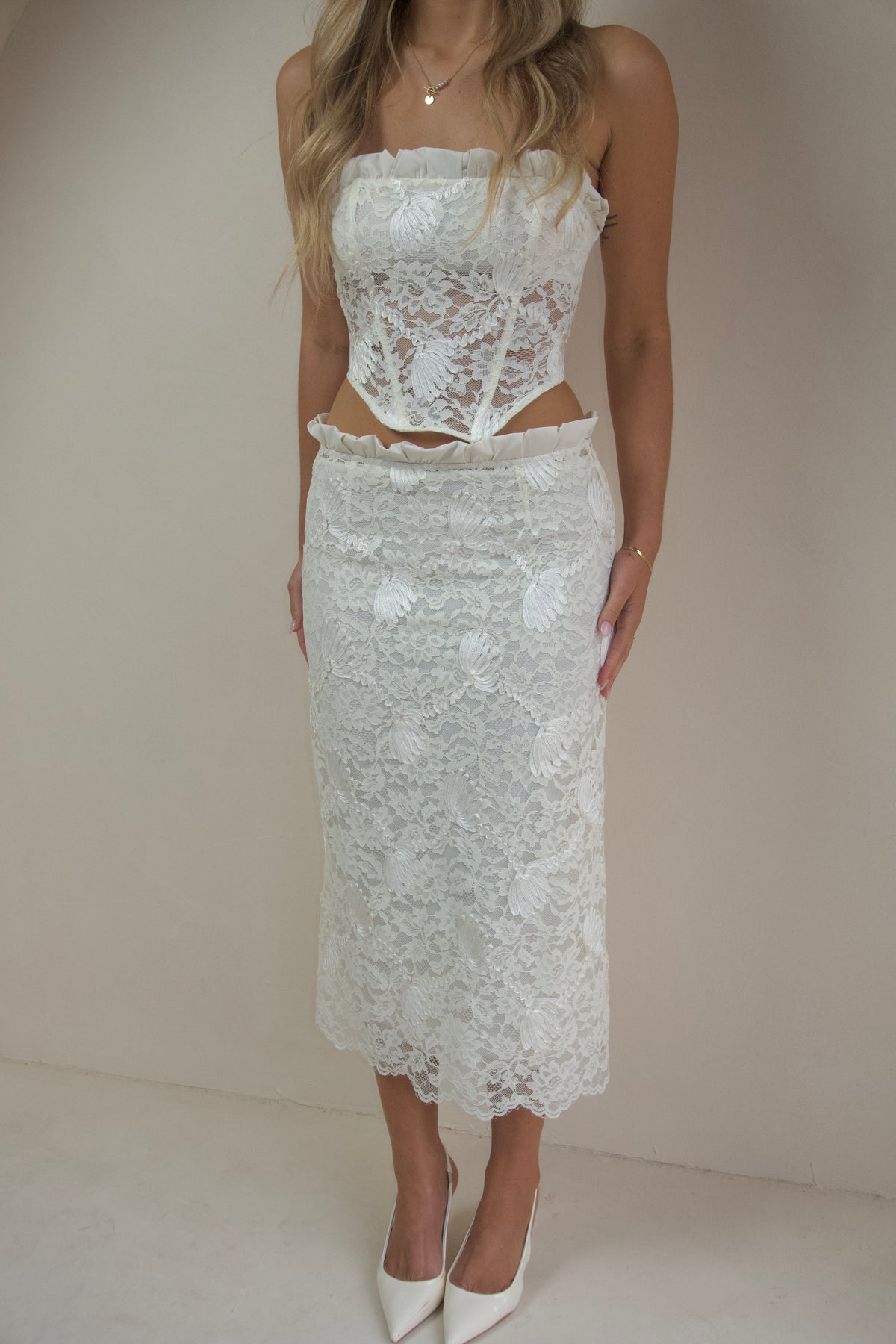  Woman wearing an ivory lace two-piece set featuring a strapless corset-style top with ruffle detailing and a matching lace midi skirt, styled with white pointed heels against a neutral studio background.