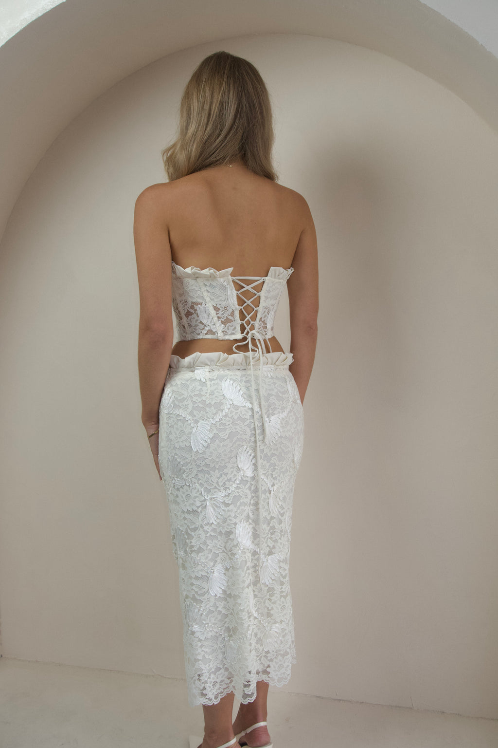 A woman viewed from behind wearing a white lace strapless tube top and matching midi skirt set, standing against a warm beige arched wall.