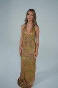 A woman modeling a sleeveless, floor-length halter gown covered in shimmering gold sequins with a subtle pink and orange floral pattern, set against a plain light gray studio background. - @lilysulfaro