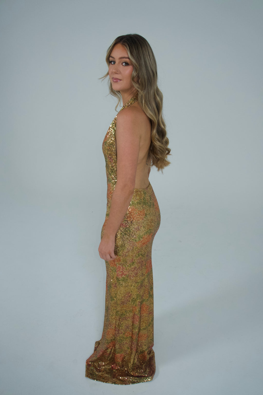 A woman modeling a floor-length, backless halter gown covered in shimmering gold sequins and metallic floral detailing, shown against a plain light gray studio background.