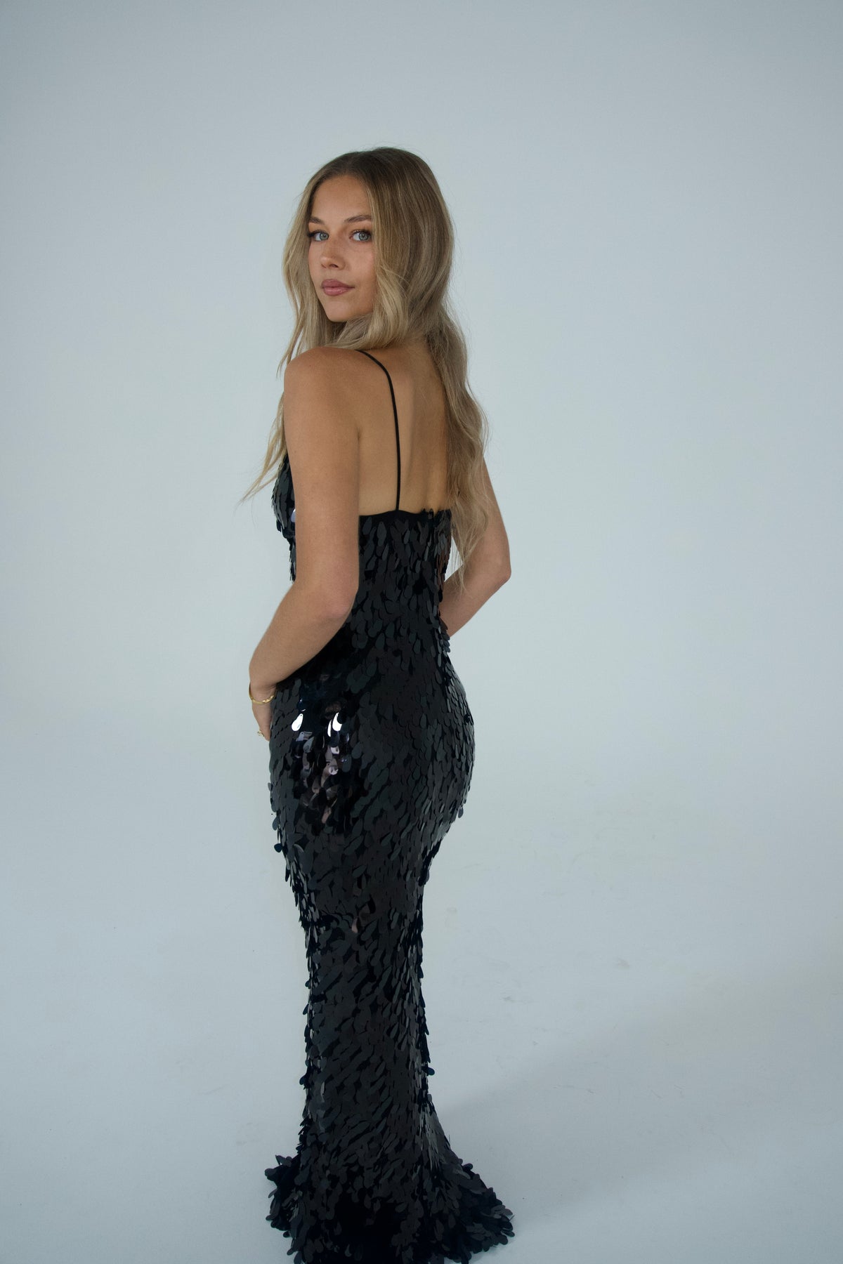 Woman wearing a black floor-length sequin gown with thin straps and a plunging V-neckline, photographed against a light grey studio background.