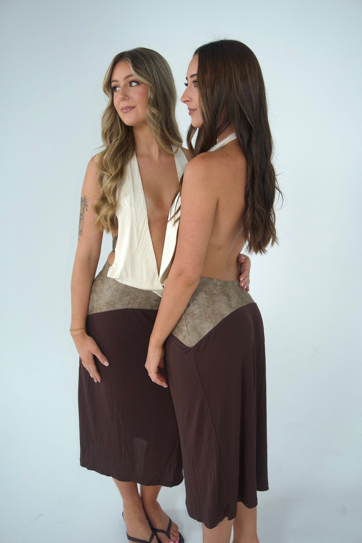 Two women modelling matching white plunging halter tops with open-back detailing, taupe textured waist panels and chocolate brown midi skirts, photographed together from a side angle against a light grey studio backdrop.