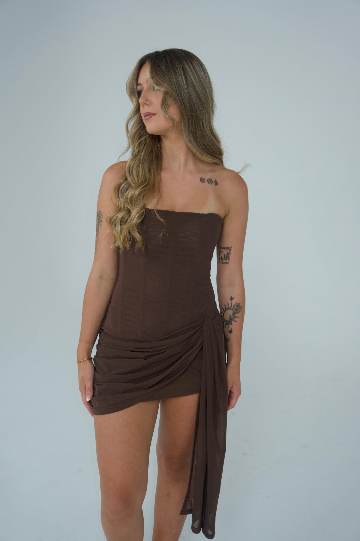 A woman modelling a chocolate brown strapless mini dress with a fitted bodice, draped wrap skirt detailing and a long cascading side sash, photographed against a light grey studio backdrop.