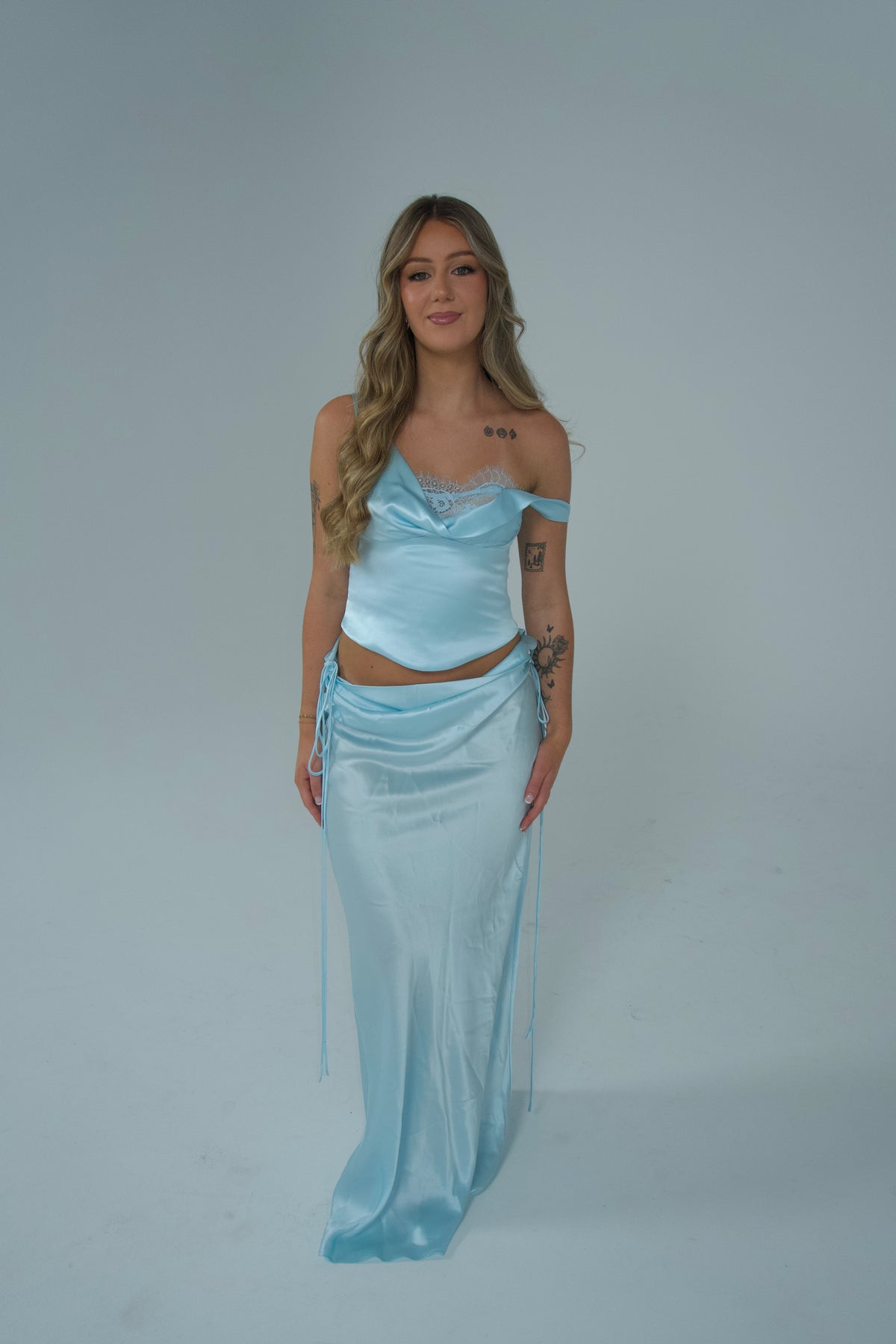 A woman modeling a light blue satin two-piece outfit consisting of an off-the-shoulder crop top and a matching long skirt, set against a plain light gray studio background.
