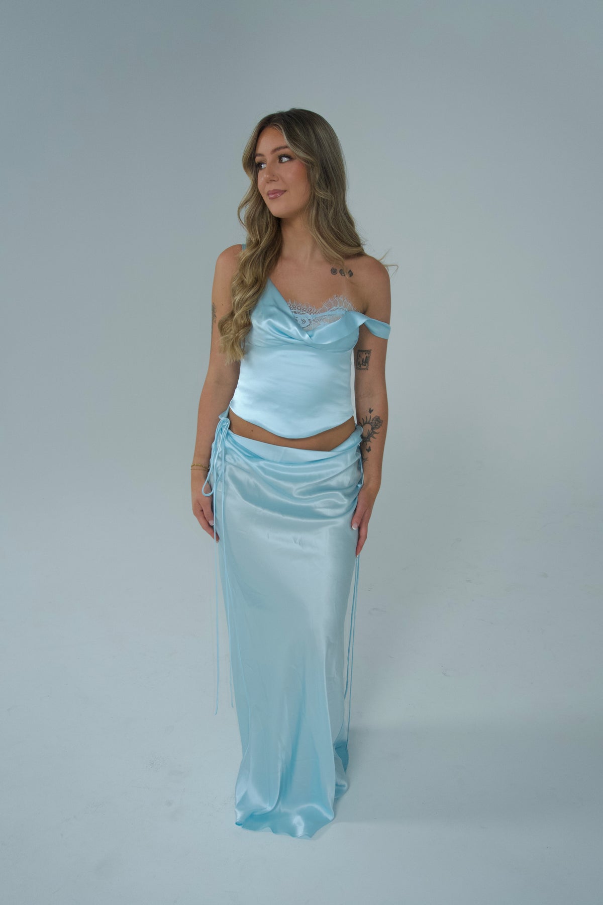 Woman wearing a light blue satin two-piece set featuring an asymmetrical lace-trim cami top and matching low-rise maxi skirt with ruched tie detailing, photographed against a soft grey studio background. - @lilysulfaro