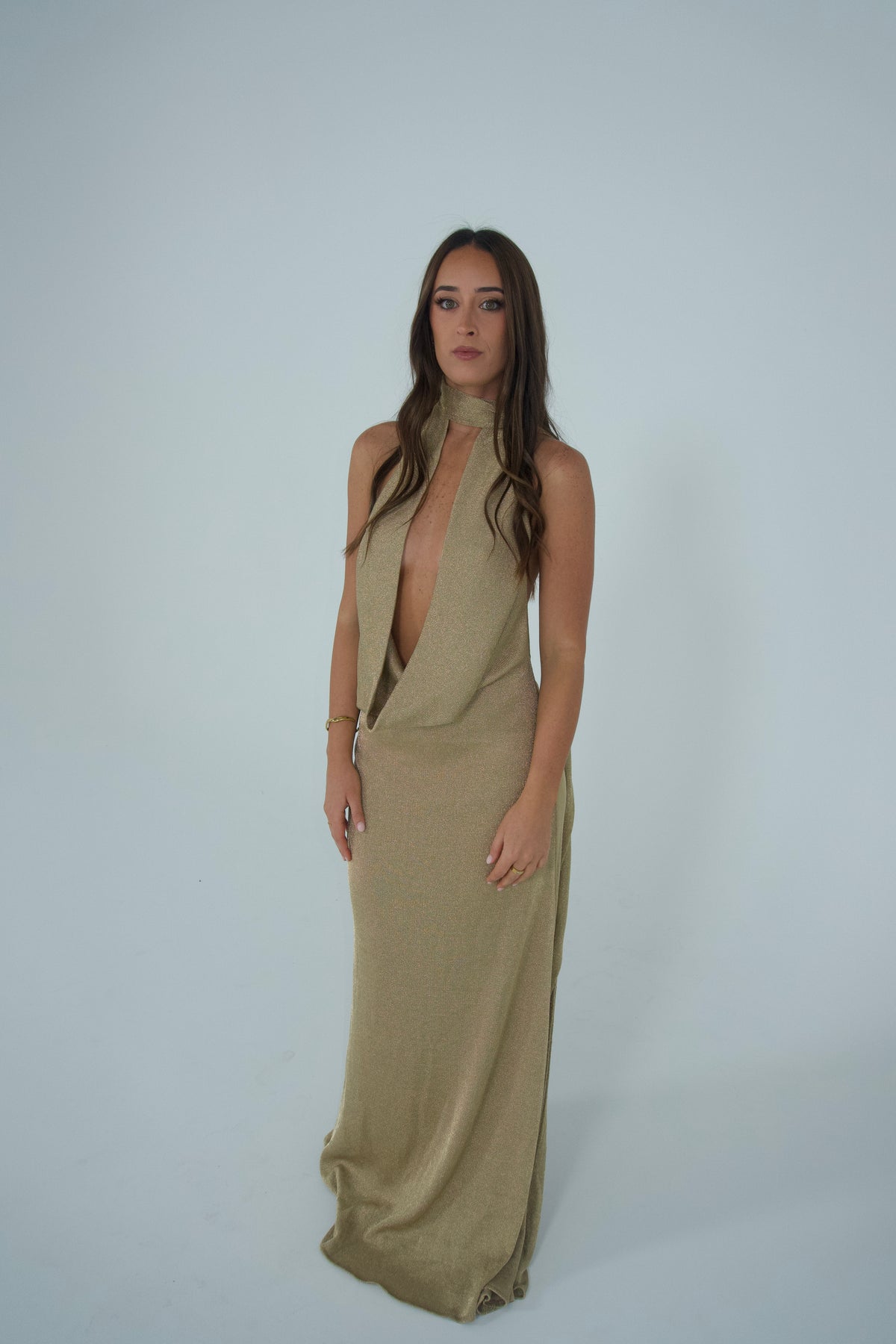 A woman modeling a sleeveless, high-neck, floor-length dress in a muted khaki-beige color, standing against a plain light gray studio background.