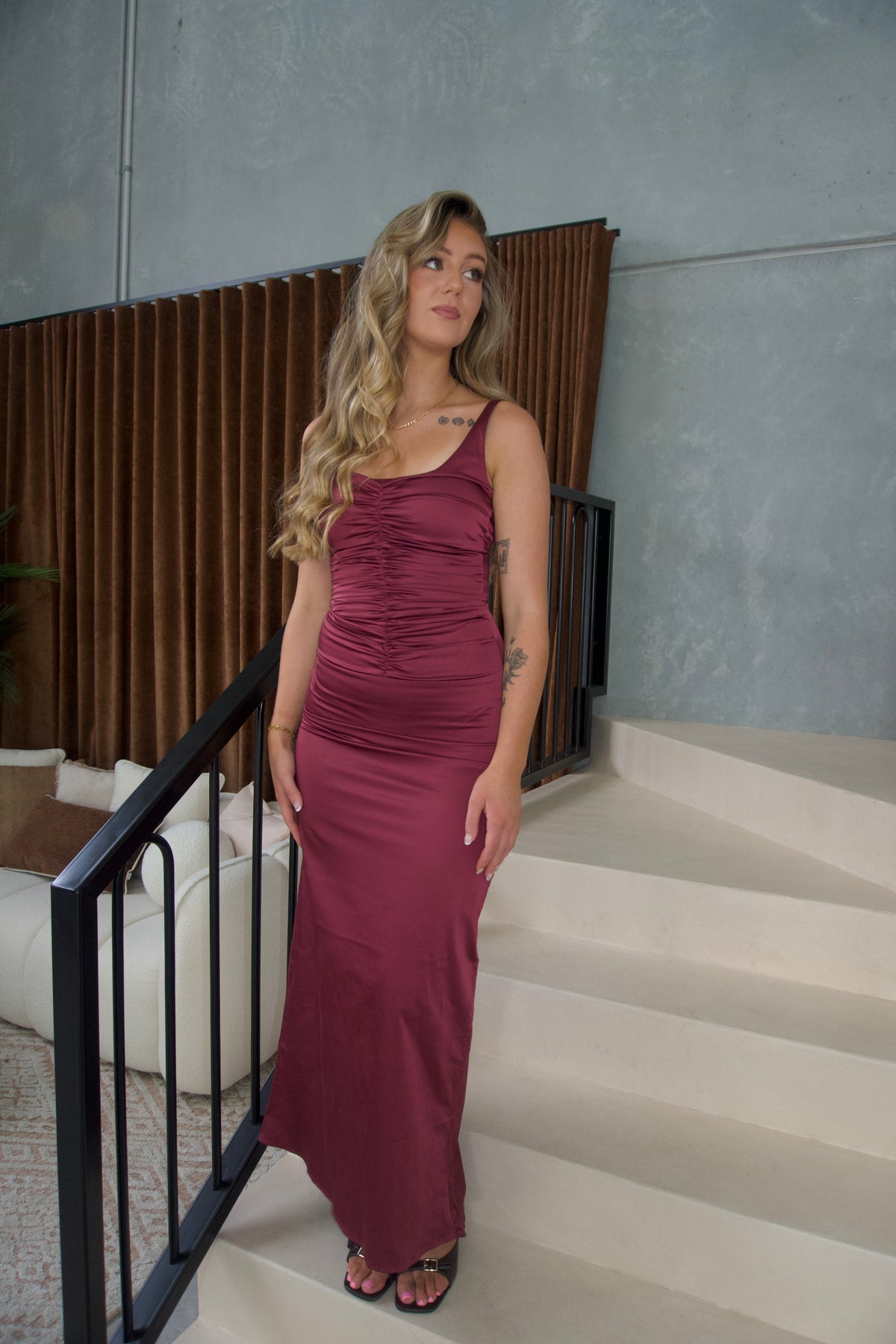 A woman with blonde hair stands on an indoor staircase wearing a sleeveless, ruched maxi dress in a deep burgundy or plum shade. The dress has a scoop neckline and a fitted silhouette.