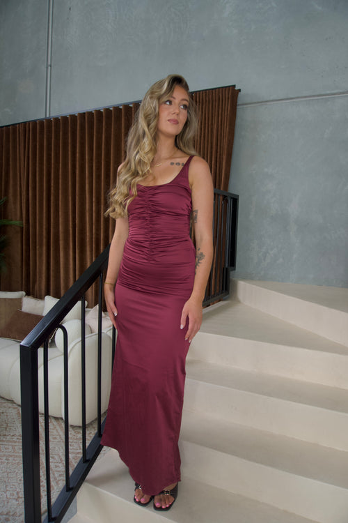 A woman with blonde hair stands on an indoor staircase wearing a sleeveless, ruched maxi dress in a deep burgundy or plum shade. The dress has a scoop neckline and a fitted silhouette.