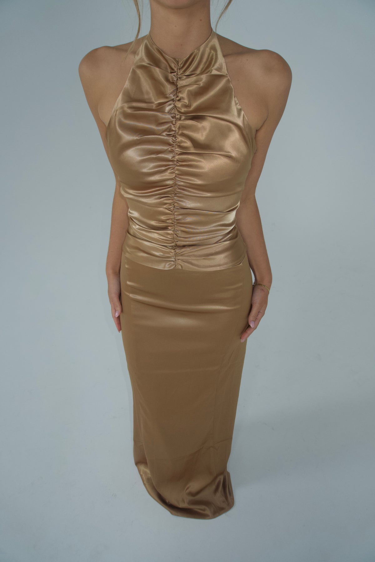 A close-up of a gold satin halter maxi dress featuring a high neckline, gathered ruching through the centre bodice and a sleek fitted floor-length skirt, photographed against a light grey studio backdrop.
