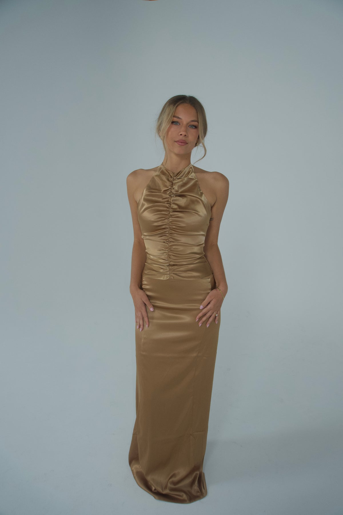 A woman modelling a gold satin halter maxi dress with a ruched fitted bodice and sleek floor-length skirt, photographed against a light grey studio backdrop.