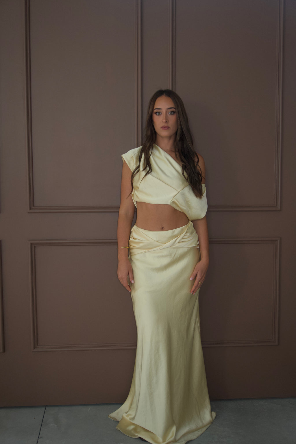 A woman modeling a pale cream satin two-piece outfit consisting of an asymmetrical draped crop top and a matching draped maxi skirt, standing against a brown paneled wall. - @sarahmadden