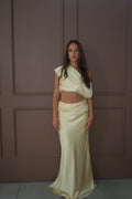 A woman modeling a pale cream satin two-piece outfit consisting of an asymmetrical draped crop top and a matching draped maxi skirt, standing against a brown paneled wall. - @sarahmadden