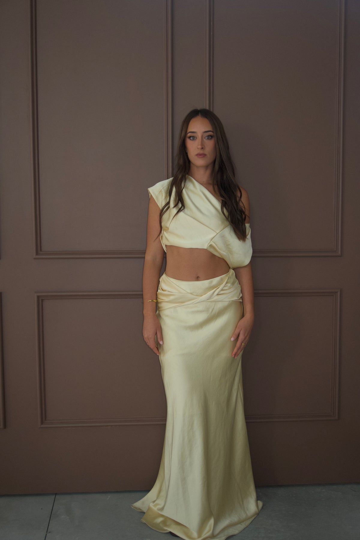 A woman modeling a pale cream satin two-piece outfit consisting of an asymmetrical draped crop top and a matching draped maxi skirt, standing against a brown paneled wall. - @sarahmadden