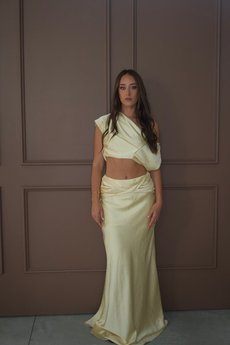 A woman modeling a pale cream satin two-piece outfit consisting of an asymmetrical draped crop top and a matching draped maxi skirt, standing against a brown paneled wall. - @sarahmadden