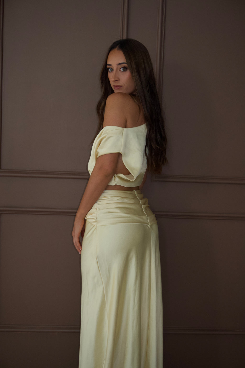 A woman wearing a cream satin two-piece outfit consisting of an off-the-shoulder crop top and a matching high-waisted pleated maxi skirt, posing against a brown paneled wall.